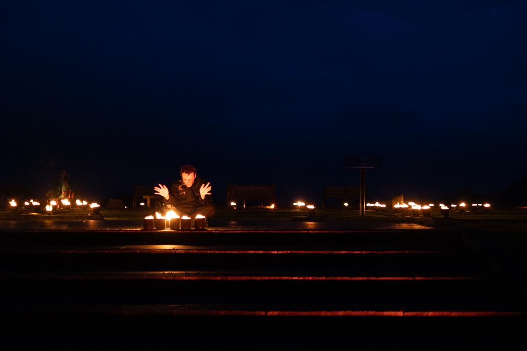 a photo of a man sat cross legged in the dark, surrounded by candles, at the top of some stairs lit by the candles. he is looking at a group of the candles, and raising his hands in a mystical way