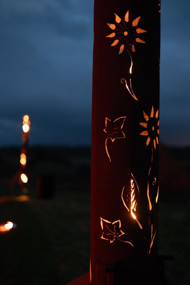 a photo of a tall metal cylinder with cutouts depicting flowers and plants. the cutouts are lit from within by fire. another cylinder is just visible in the background as a bright blur