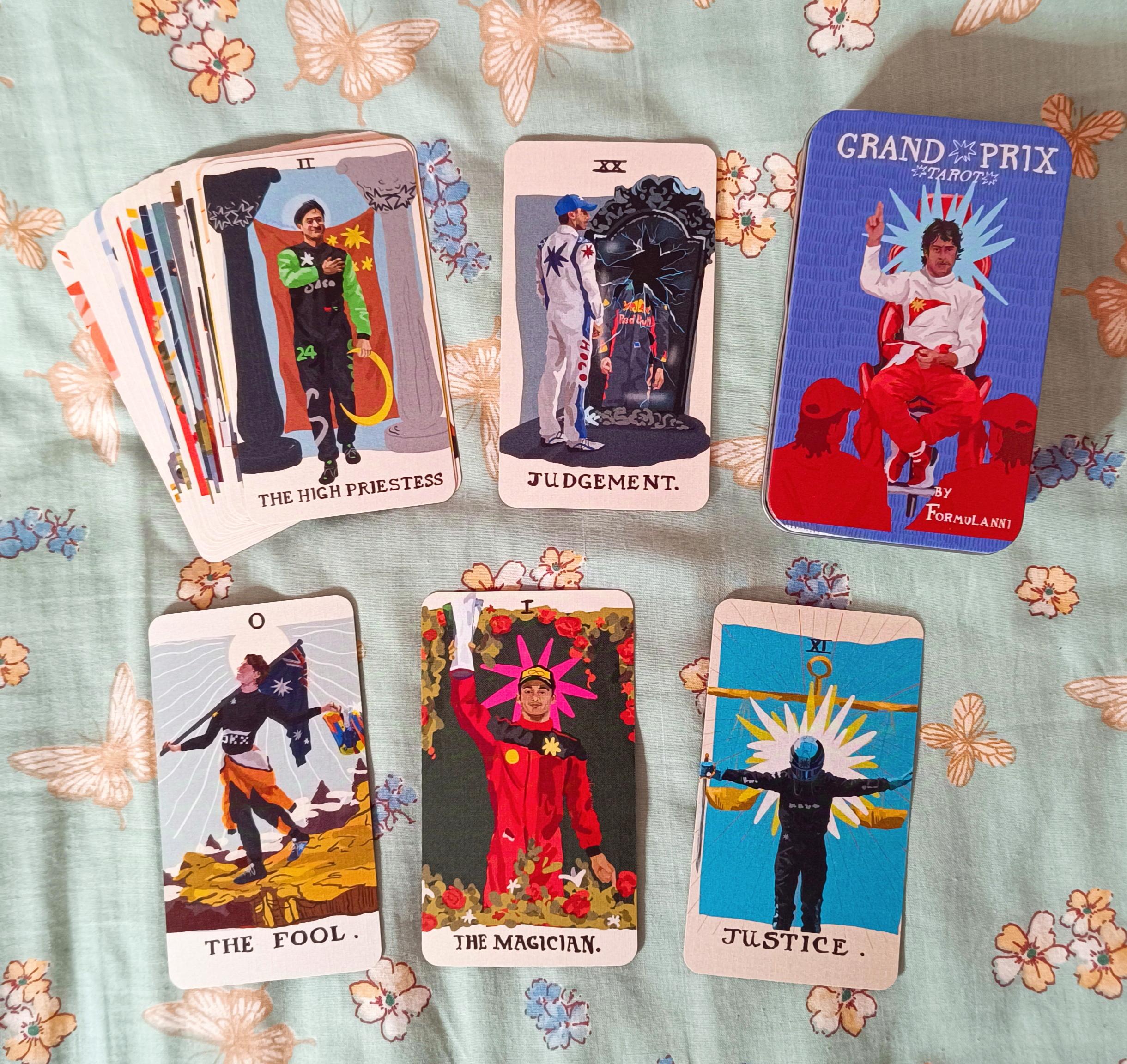 Photo of the Grand Prix Tarot deck by formulanni. The tin box features Fernando Alonso at The Hierophant. The deck is spread out a bit, and the cards that are in full view are: Zhou Guanyu as The High Priestess, Daniel Ricciardo as Judgement, Oscar Piastri as The Fool, Charles Leclerc as The Magician and George Russell as Justice.