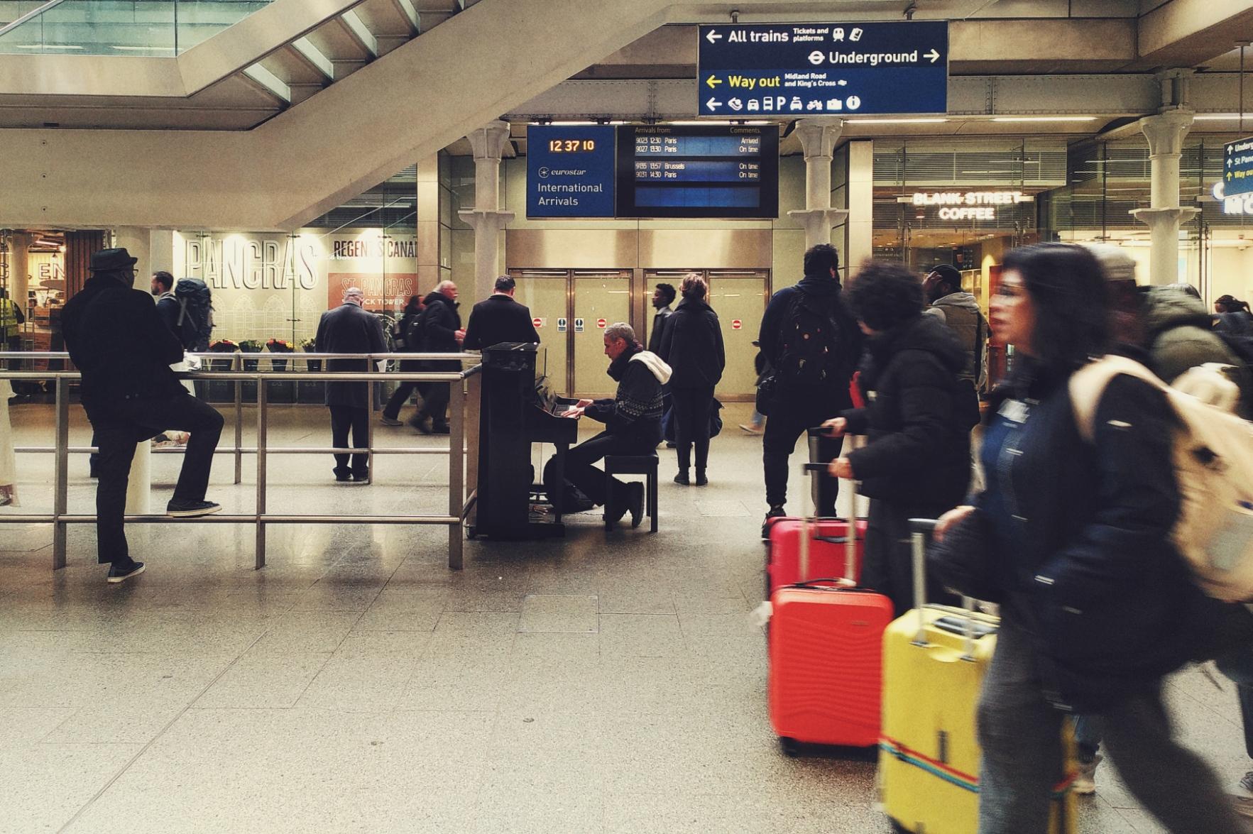 A horizontal colour photograph a train station hall. A person is playing a piano in the middle of the scene. Folks pass by with colourful suitcases, other folks stand around looking at a display screen in the background.