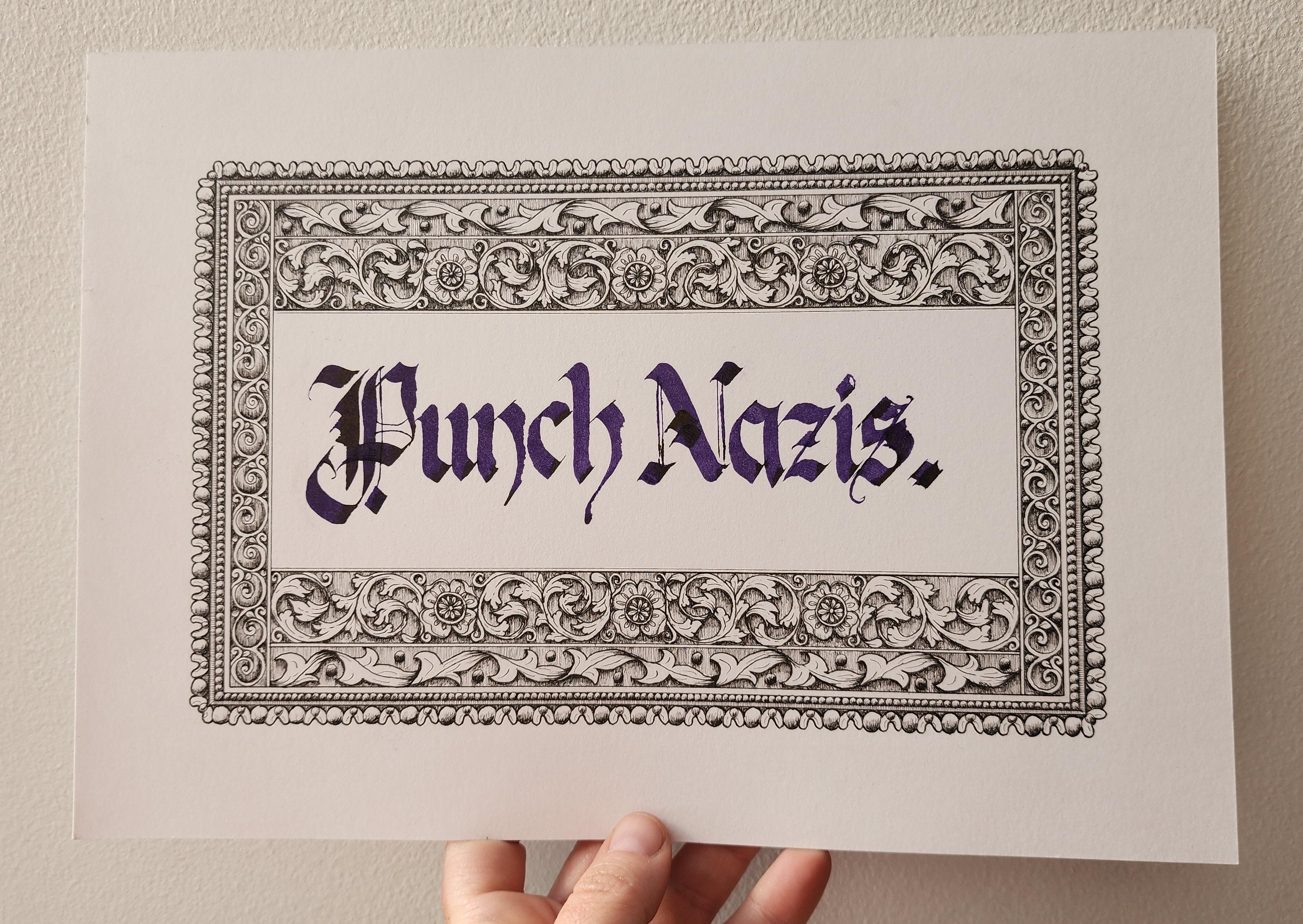 My hand holding up a piece of artwork against a white wall.
'Punch Nazis.' calligraphed in purple ink, blackletter script, and surrounded by a hand-drawn decorative border of repeating patterns of swirling leaves, flowers, and circles, shaded to look like a 3D engraving.