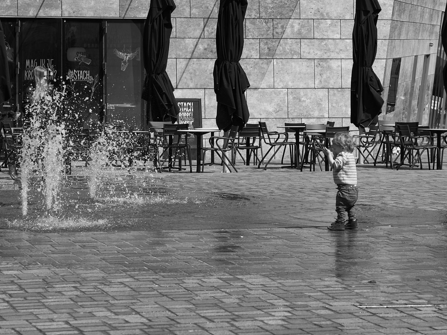 A young child is playing near a water fountain in a plaza, surrounded by outdoor café seating. The image is in black and white, capturing the moment as the water splashes around the child.