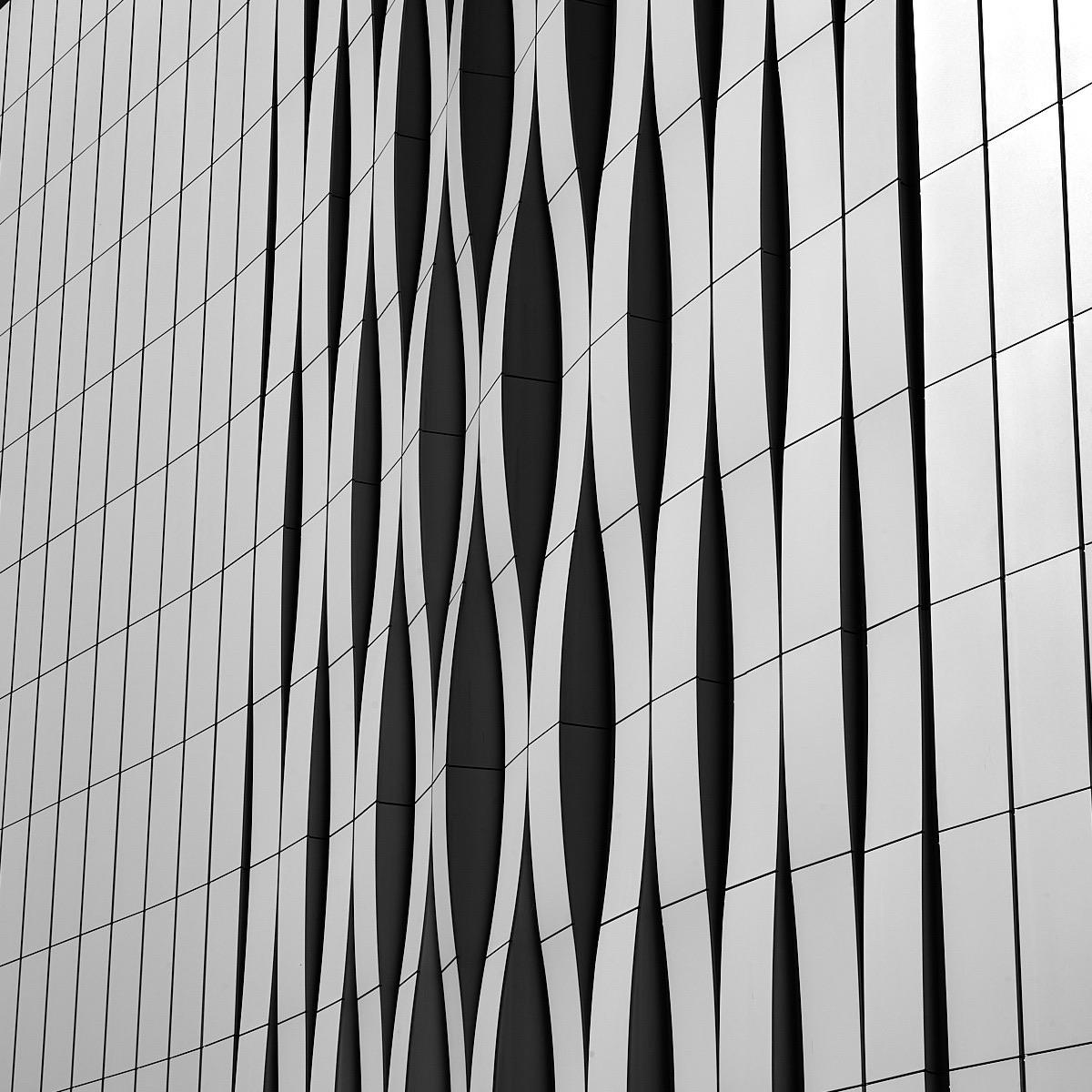 A close-up view of a modern building facade featuring dynamic wave-like patterns in a monochromatic color scheme. The design includes alternating white and gray panels that create a visually striking texture.