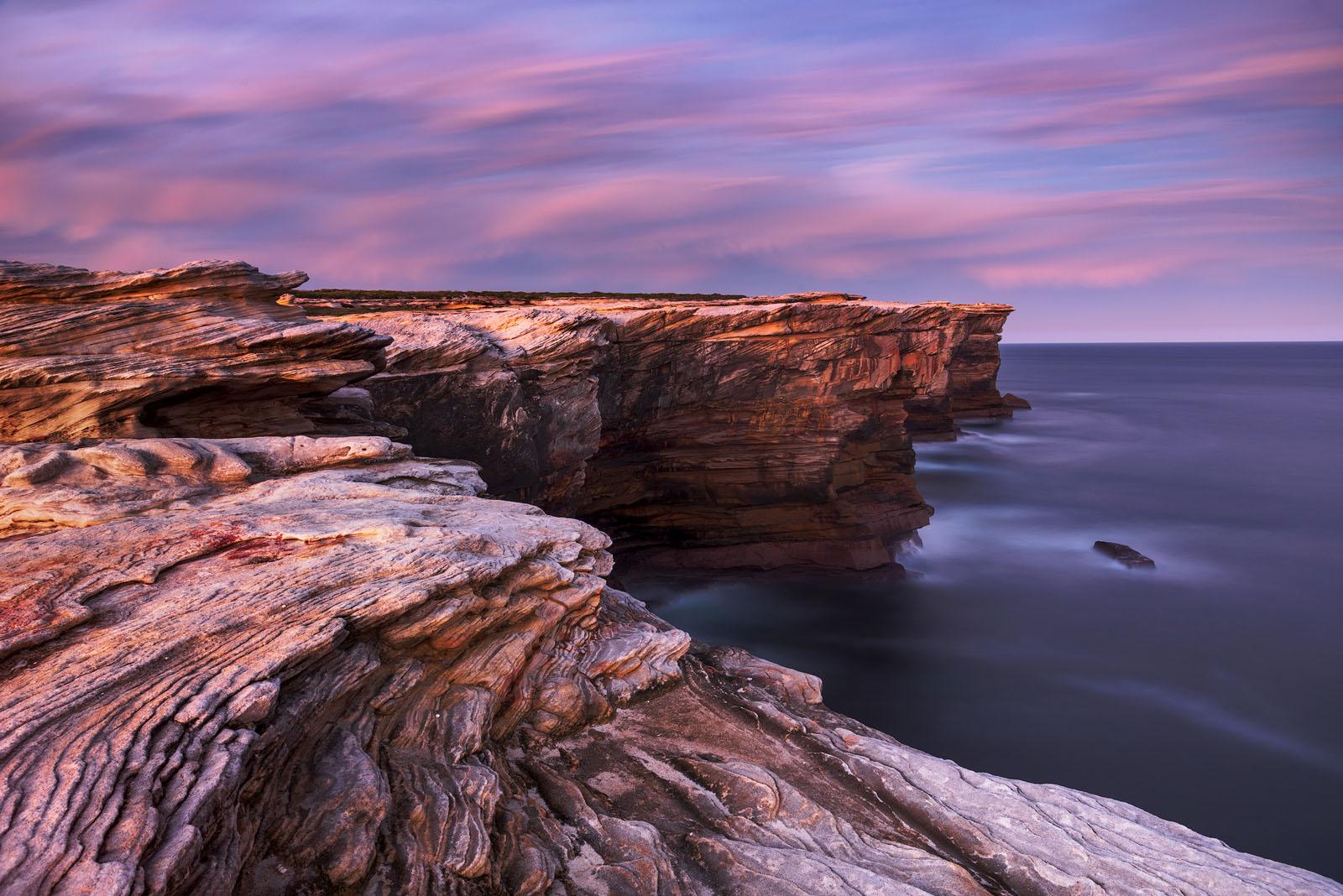sandstone cliffs facing the sea with curved patterns during the sunset. Both the sky and the cliffs are pink. The sea is smooth due to the long exposure