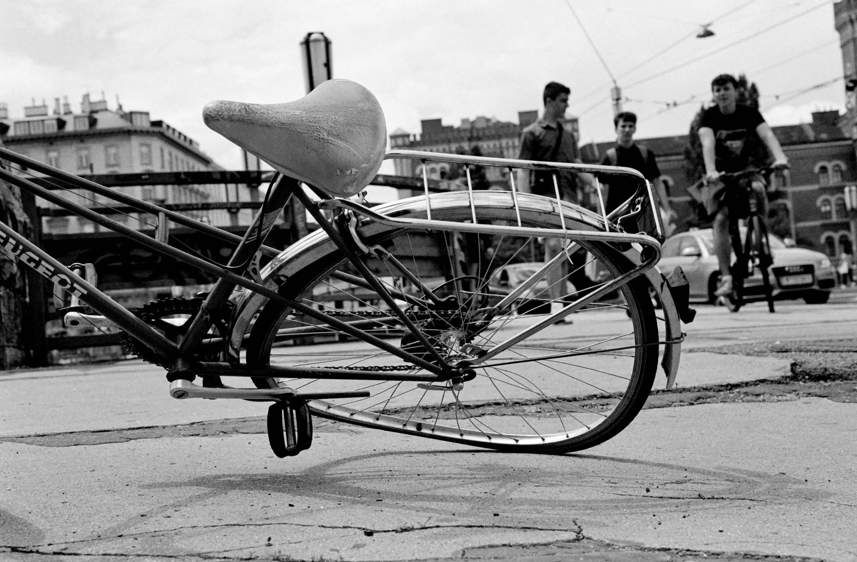 The black and white image shows a close-up of a bicycle lying on its side on a cracked sidewalk, with its rear wheel and saddle prominently in the foreground. In the background, three young men walk while another rides a bicycle toward the camera, with city buildings and cars visible behind them.