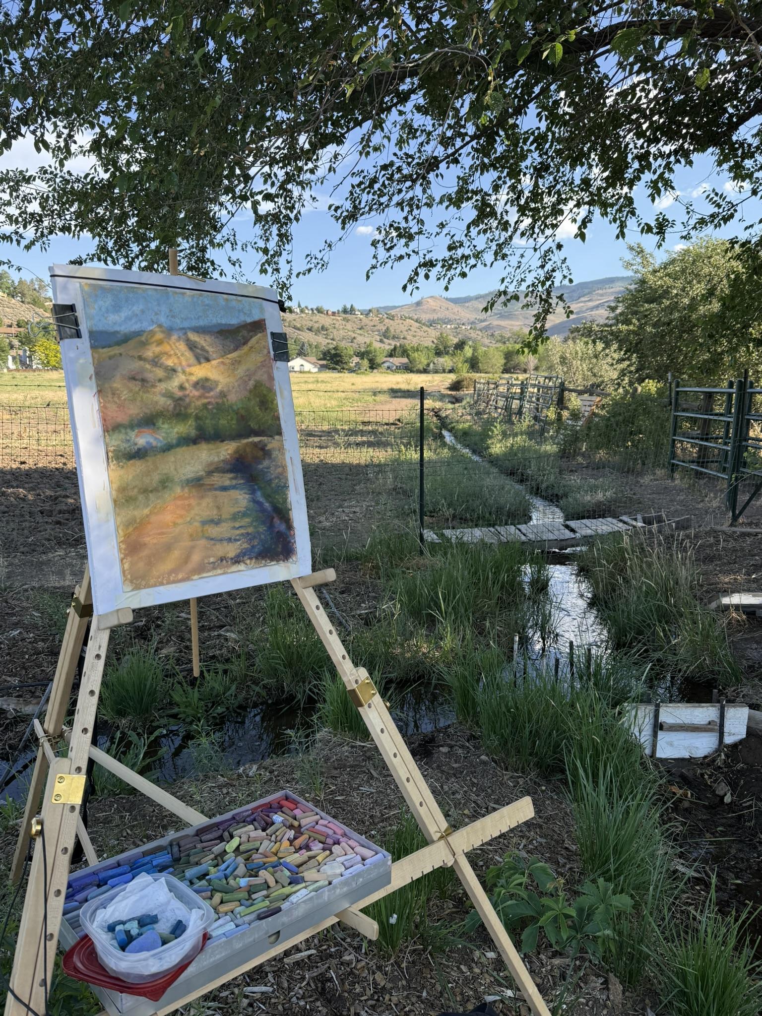 An outdoor easel is set up under a tree, and a work-in-progress pastel painting is on it. Far mountains, a meadow and a small streal is in the background.