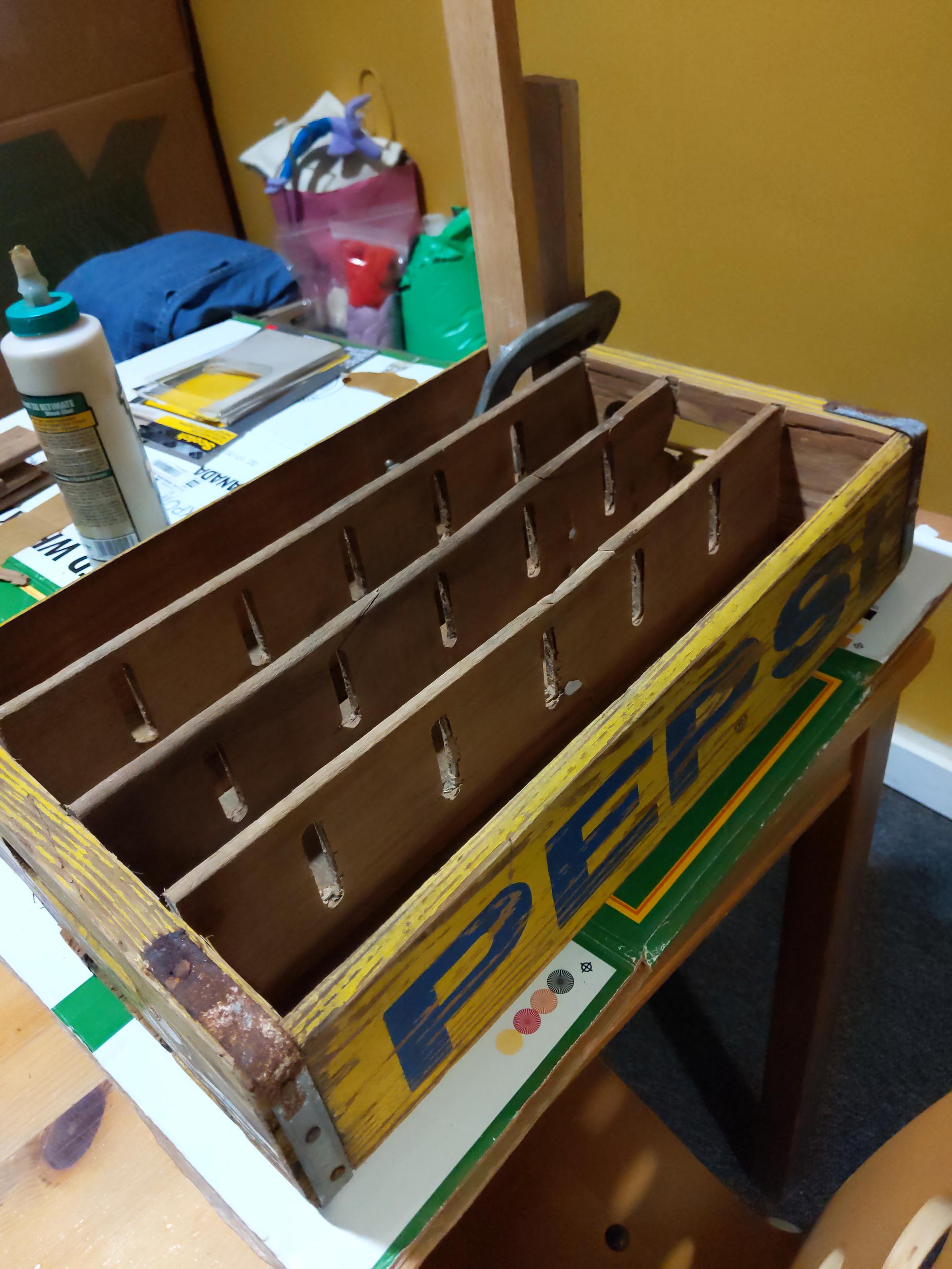 A photo of a very old pepsi branded wooden crate with clamps on one corner and half the dividers removed.
Much basement has been scrubbed off of it.