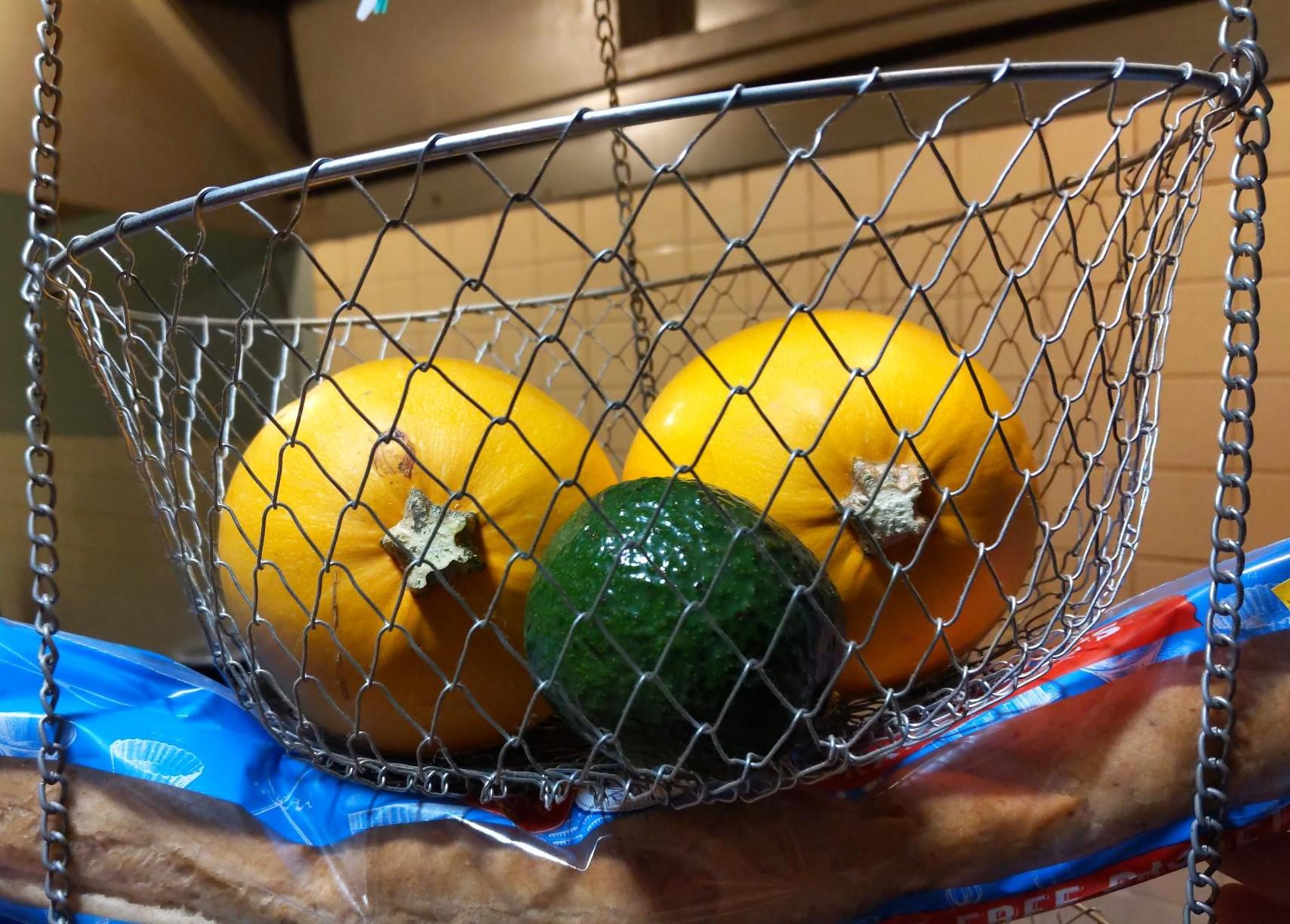 A pair of squashes, an avocado, and a baguette forming a face in a wire basket.