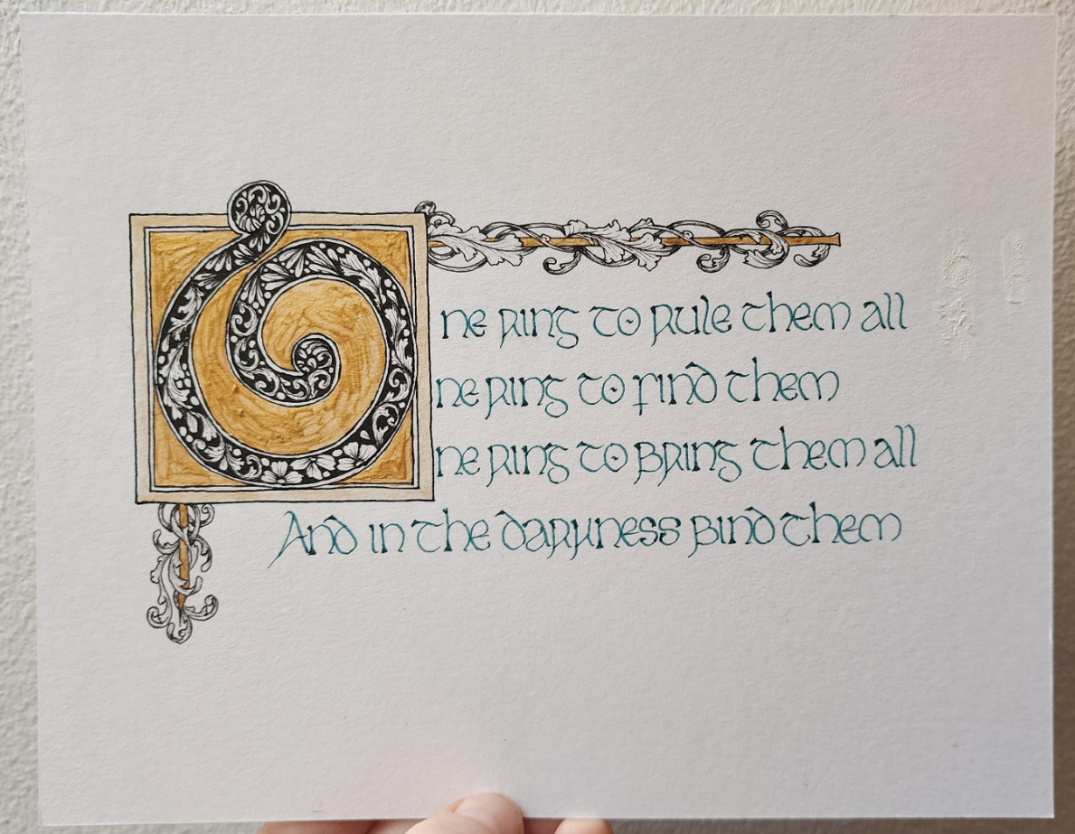 Calligraphed in teal-coloured ink in insular script, and with a big ornate 'O' filled with black ink patterns and surrounded by gold;
'One ring to rule them all
One ring to find them
One ring to bringthem all
And in the darkness bind them'
The box that the 'O' is in has bars extending from the top right, and bottom left, wrapped in black ink line art curling vines.