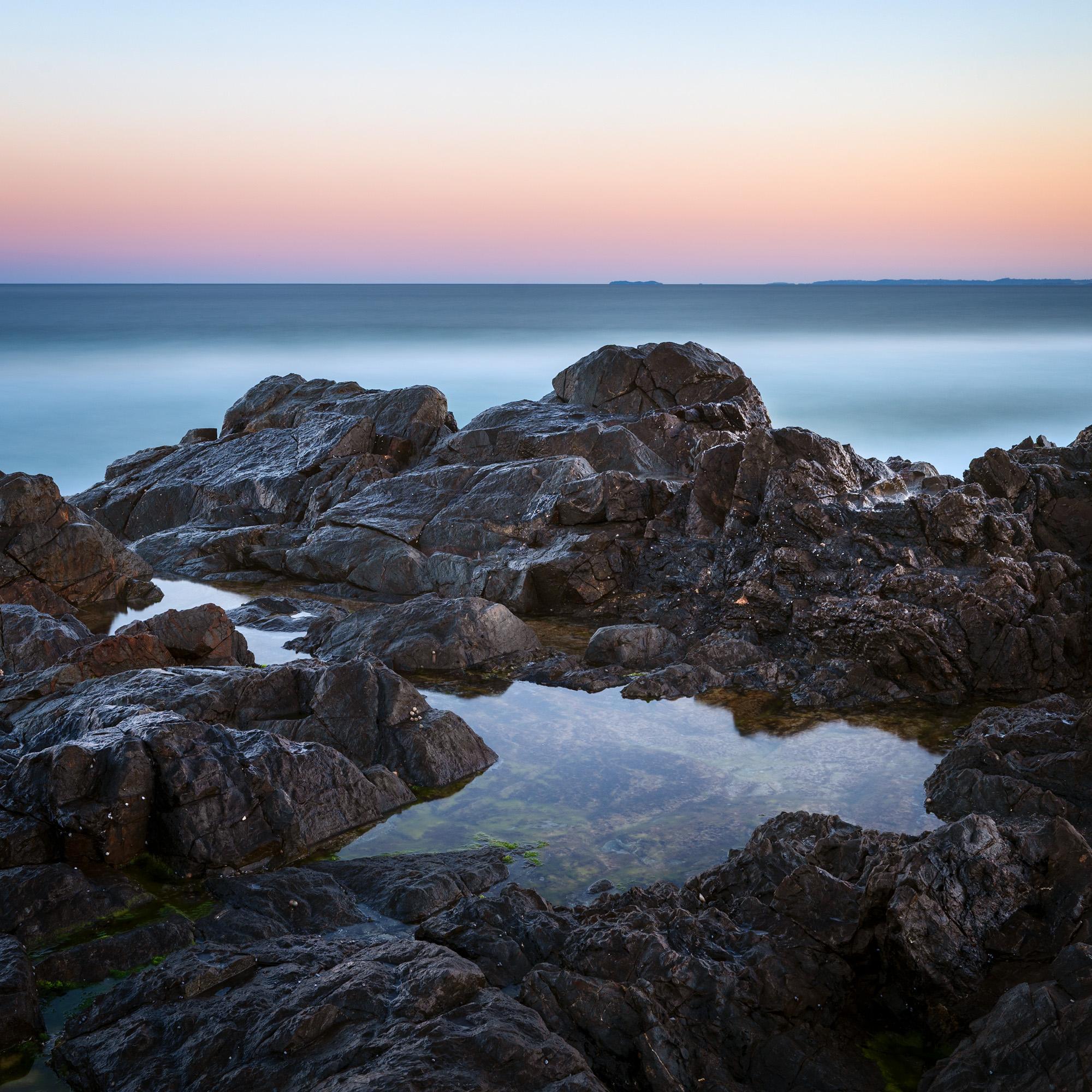 a serene evening seascape with rocks around a small still puddle, smooth sea and a Venus belt sky