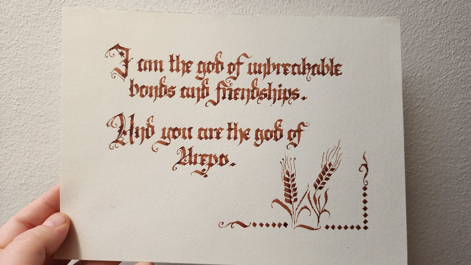 Calligraphed in a fancy gothic embellished blackletter script in red-brown ink;
"I am the god of unbreakable bonds and friendships. And you are the god of Arepo."
There's an illustration of two wheat stalks in the corner, drawn with the calligraphy pen, with some other simple pen-made decorations going along the bottom and up the side of the page a little way.