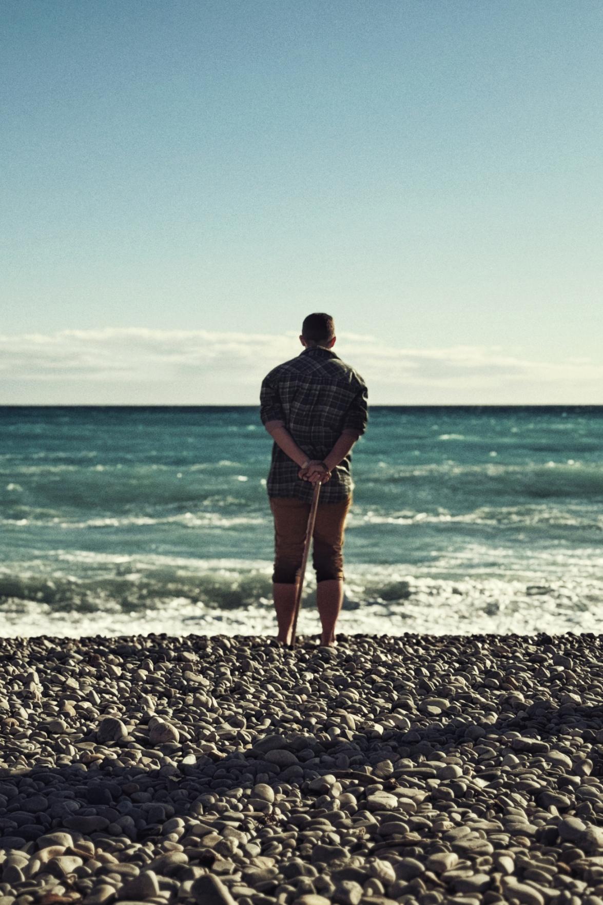 A vertical colour photograph of me looking at the sea. The sea is blue and a little frothy. The beach is all grey rounded pebbles. I am in the middle, mustard jeans rolled up to my knees. They are damp at the bottom edge. I have a blue and green and yellow checked shirt. My hands are behind my back holding a wooden stick. I have short dark cropped hair.