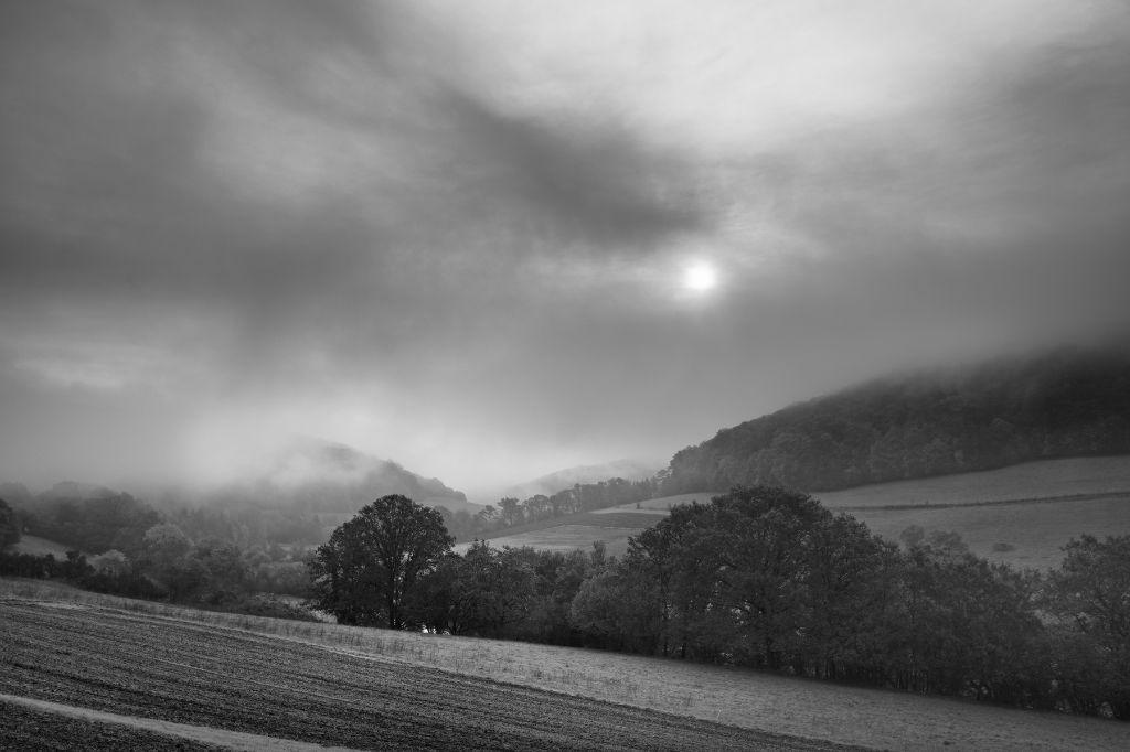 Monochrome landscape photo of the morning sun fightung bravely against low hang fog and clouds. A veil is lifted from the hills and woodlands and a new day begins