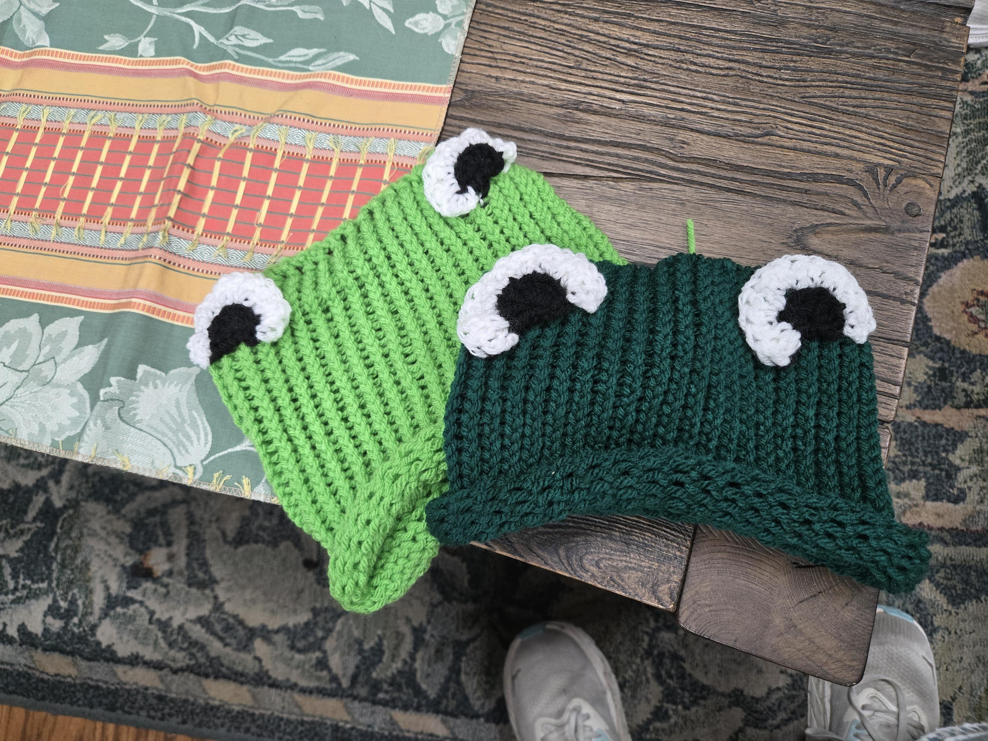 Two green knitted hats on a table. Both are square shaped so that the two corners will stick up when on your head. The eyes are a black half circle surrounded by a white trim, giving the eyes a cartoon look.