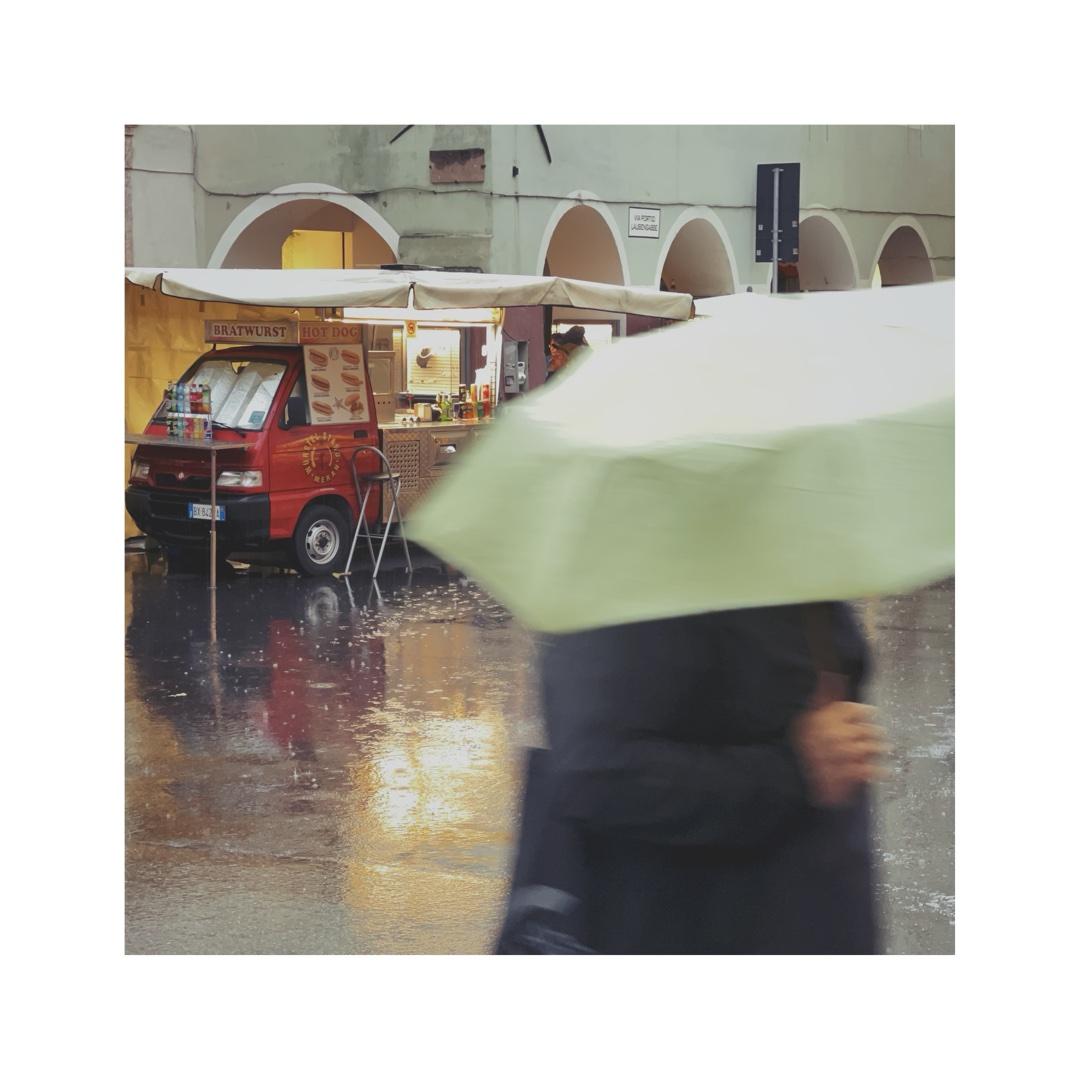 a square street image. it's raining, people are walking in the streets with their colorful umbrellas. in the foreground there's one of them, slightly blurry, walking with a green umbrella covering his face. in the background there's a little van selling hot dogs. its lights are being reflected on the wet street
