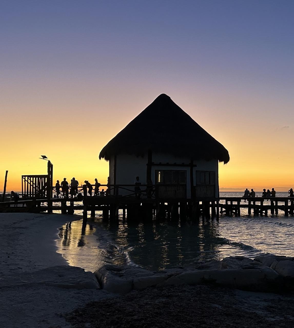 Against a sunset gradient sky if grey, blue, mauve, orange, and bright yellow are silhouettes of a wooden pier with many people on it & a round hut with pointed roof built on the pier over the water. In the foreground, the sunset colors are reflected in water with small waves rolling in, and sand and rocks.