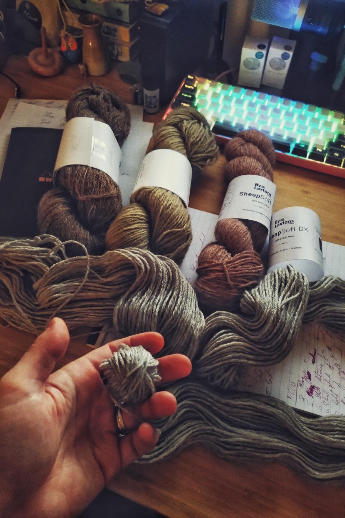 A vertical colour photograph of four skeins of wool on a desk by a little up keyboard. One is brown, another is green, another is pink. The last one, laid open across the desk is grey. My hand, at the bottom left of the image holds the start of a handmade ball of yarn.