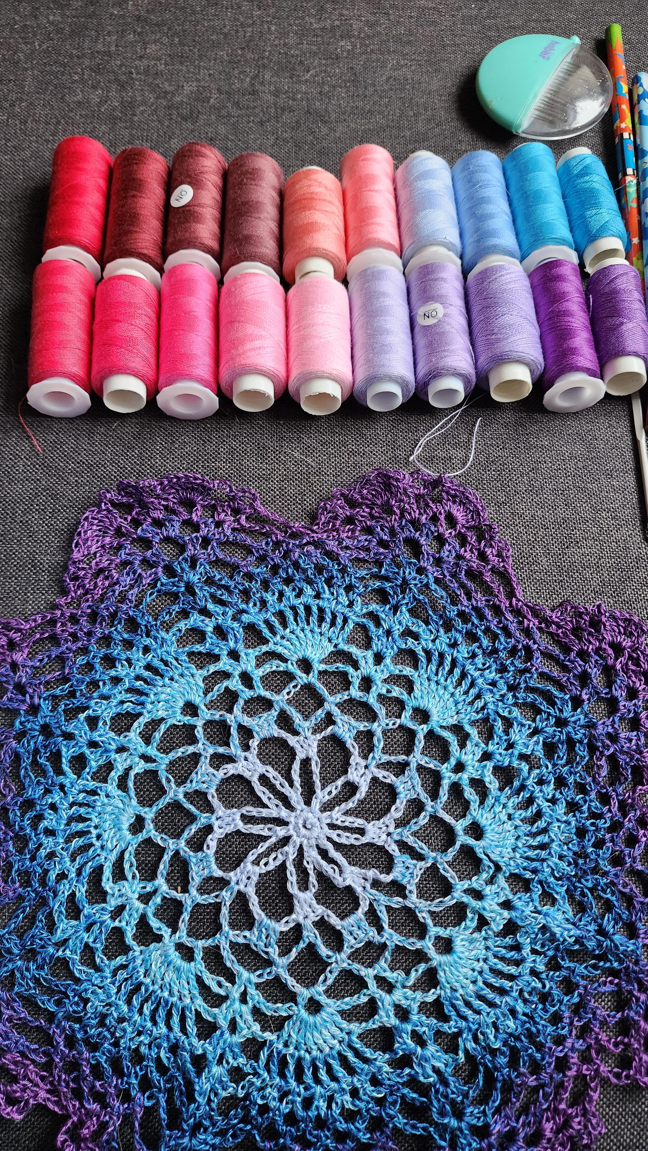 A crocheted doily in blues and purples, with spools of thread laid out above it graduating through purple to pinks, reddish browns, and into blues