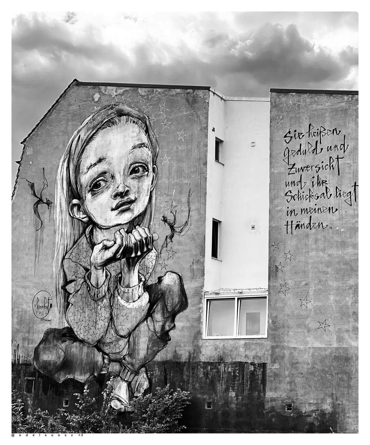 Aufnahme in schwarzweiß, Giebelseite eines Altbauhauses, dazwischen mehrere kleine Fenster, auf der großen hellen Fläche eine riesiges Graffiti, dargestellt ist ein im Schneidersitz sitzendes kleines Mädchen mit bettelten Händen, auf der Fläche daneben der ausgeschriebene Spruch: „Sie heißen Geduld und Zuversicht und ihr Schicksal liegt in meinen Händen.“