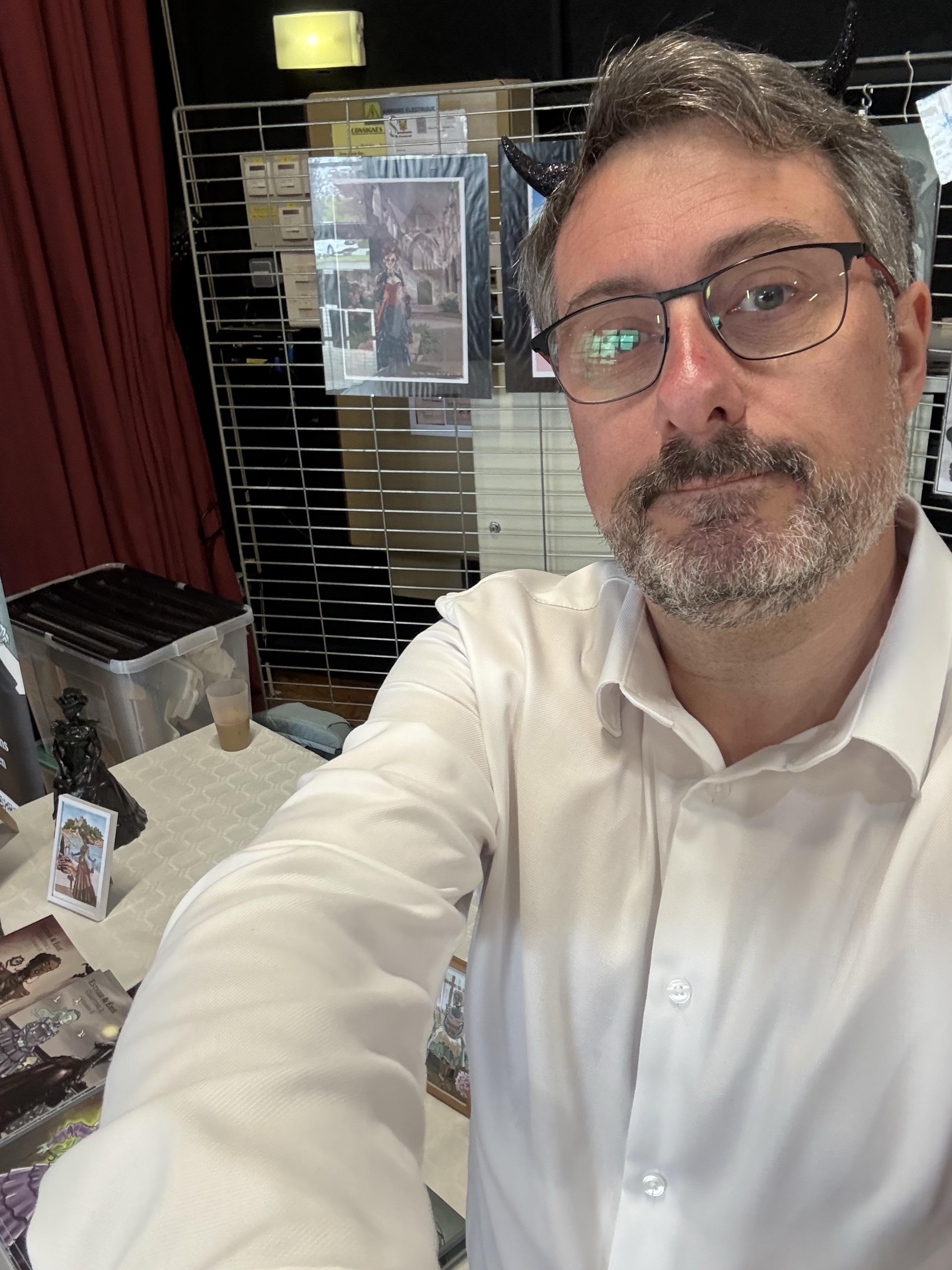 Me taking a selfie in a setting featuring artworks displayed on a grid backdrop. He is wearing a white shirt and has novelty horns. In the foreground, there are items on a table, including books, a small figurine