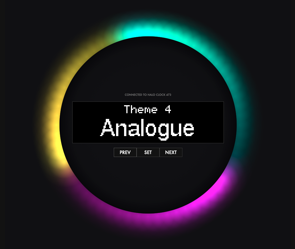 The image shows a circular digital interface with a glowing halo effect in multiple colours—cyan, yellow, and purple—around its perimeter. At the centre of the circle, there's a digital display with the text "Theme 4 Analogue" in a pixelated font. Above this text, it reads "CONNECTED TO HALO CLOCK 473." Below the main text are three rectangular buttons labelled "PREV," "SET," and "NEXT," suggesting navigation options for selecting different themes. The background is dark, which accentuates the vibrant halo colours.