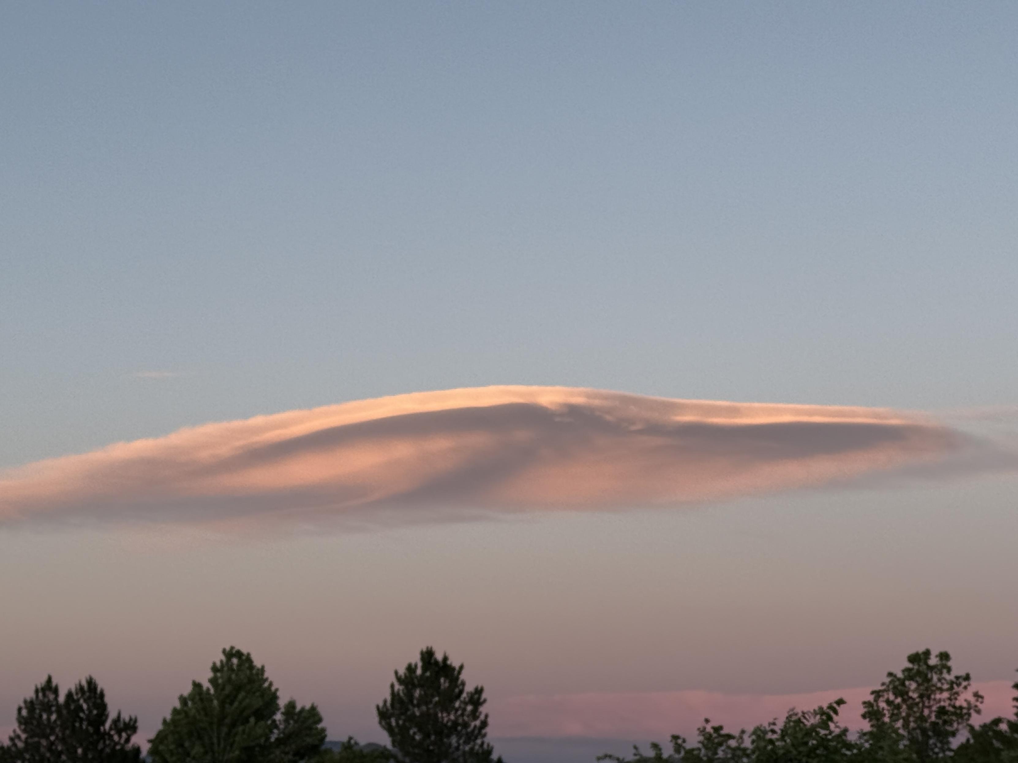 The setting sun is illuminating a giant wave cloud into orangy pink and violet above the tree line.