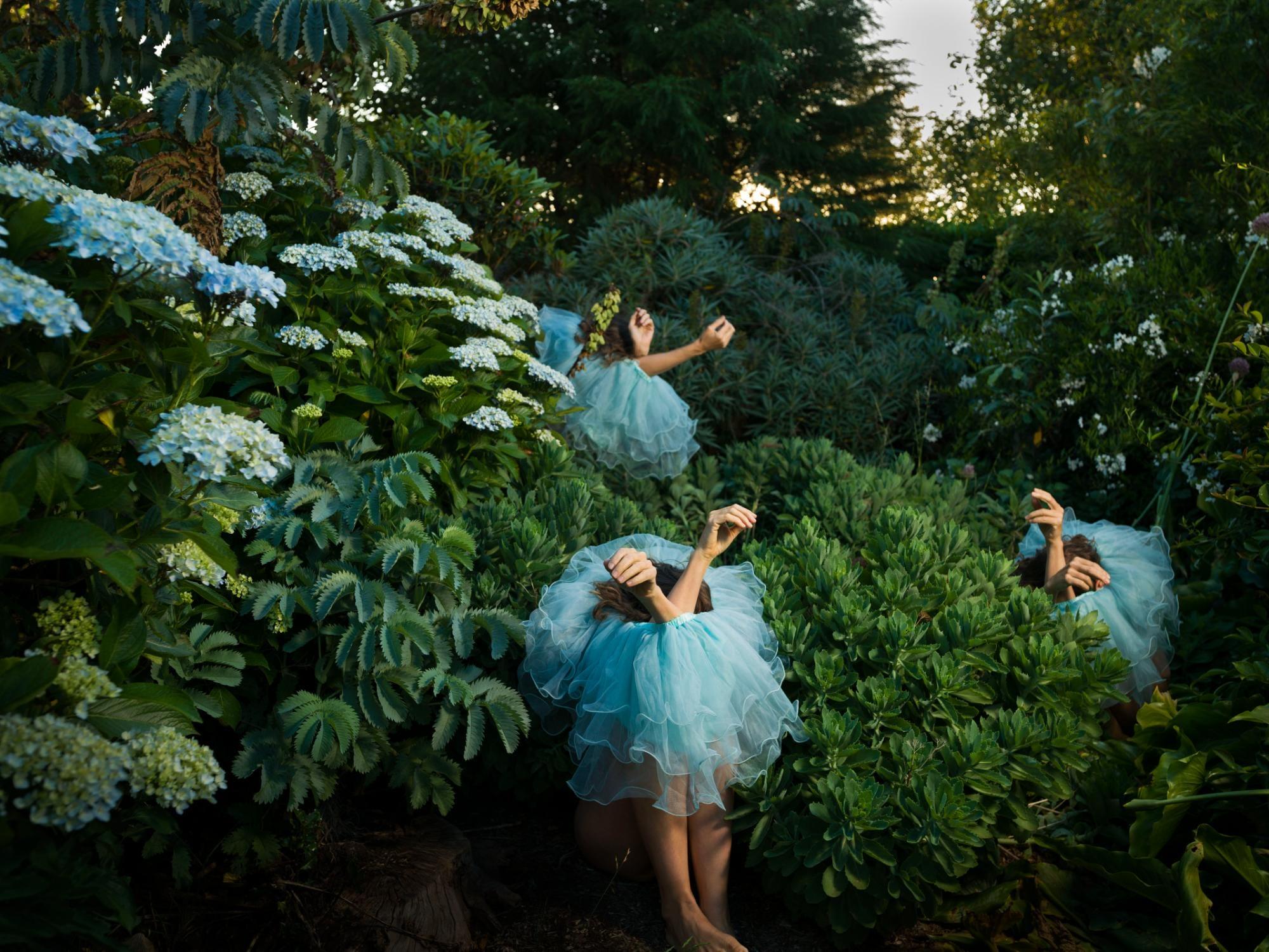 a photograph by Tamara Dean of three individuals sitting in a dense brush, wearing blue tutus around their bodies, only their arms sticking out