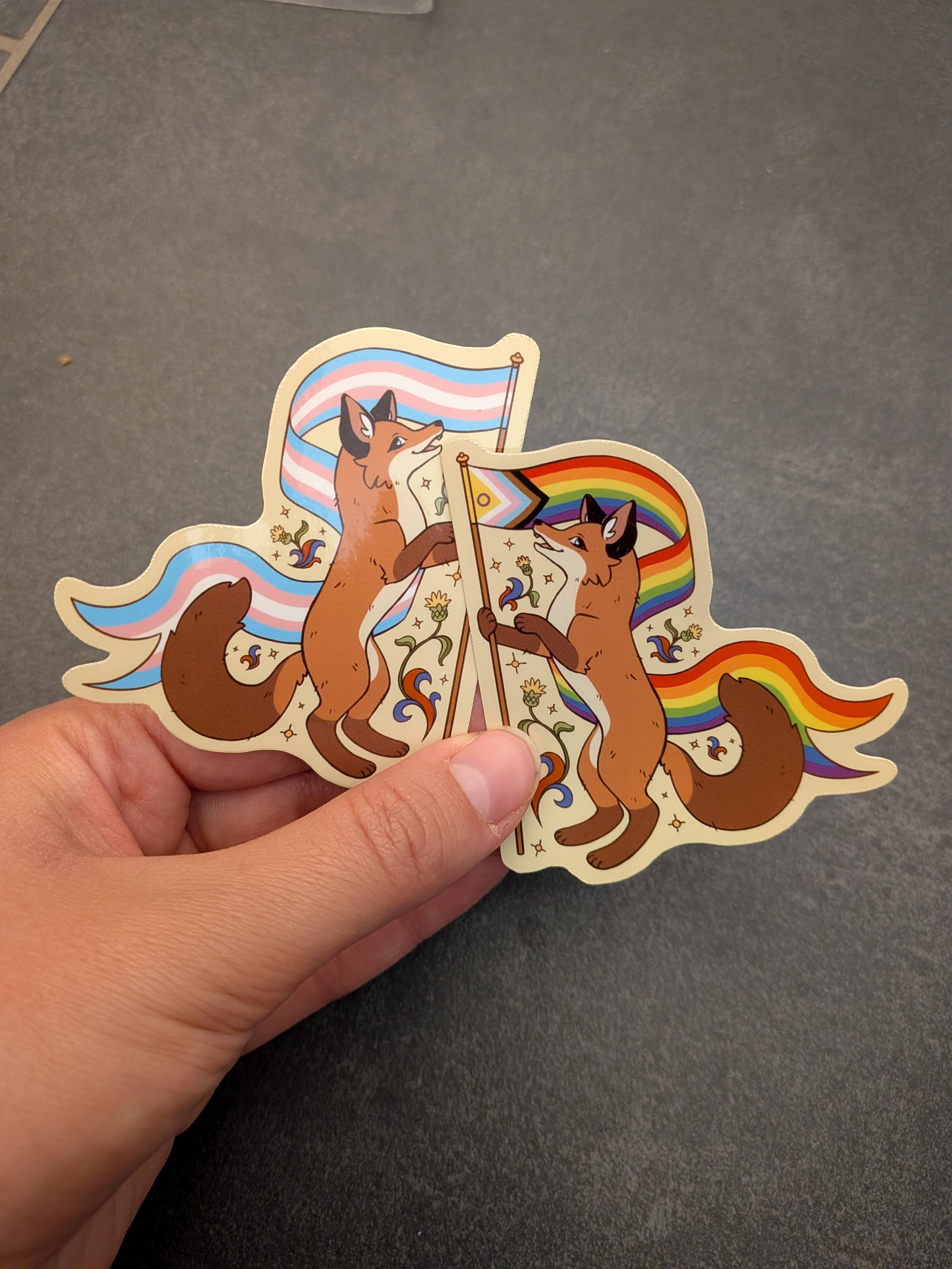 Two stickers with foxes each holding a pride flag. One is a trans flag and the other is a rainbow flag.