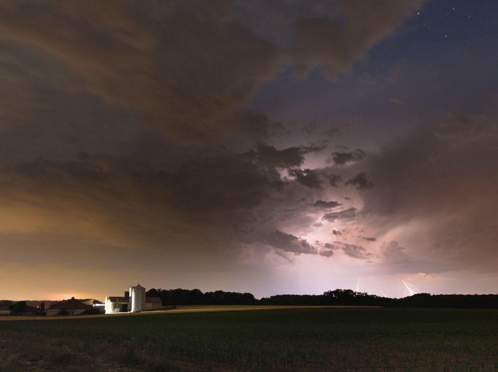 Photo of a nightly landscape. Clouds are lit up by lightning. A small farm calmly standing by off to the side