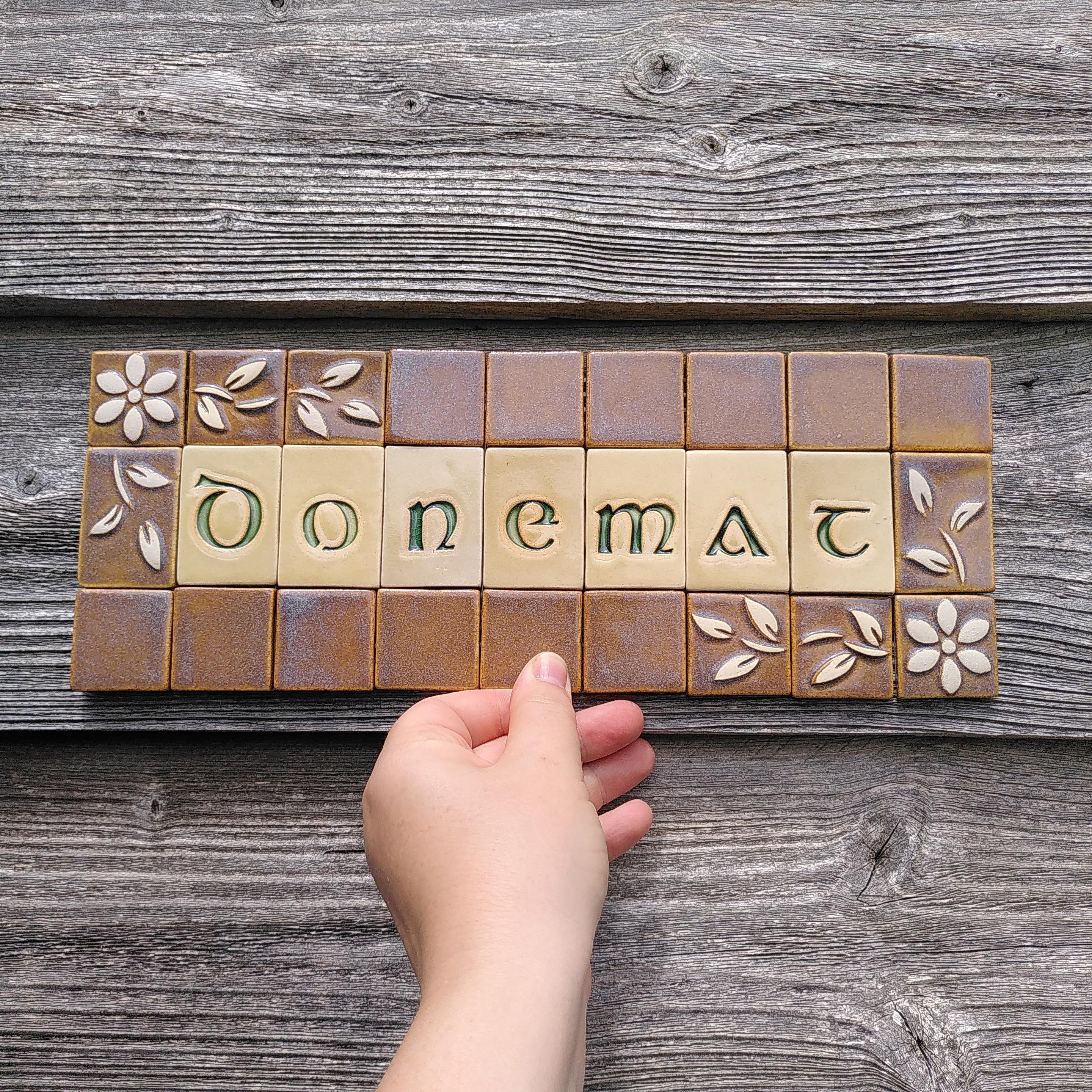Une plaque en mosaïque en céramique. Composée de 3 lignes de carreaux et rectangles. Au milieu est écrit Donemat en breton (bienvenue) en lettres estampées vert émeraude sur fond beige. Une lettre par pièce. Le contour est composé de carreaux brun/bleu avec dans deux coins opposés des fleurs et feuilles blanches en relief.