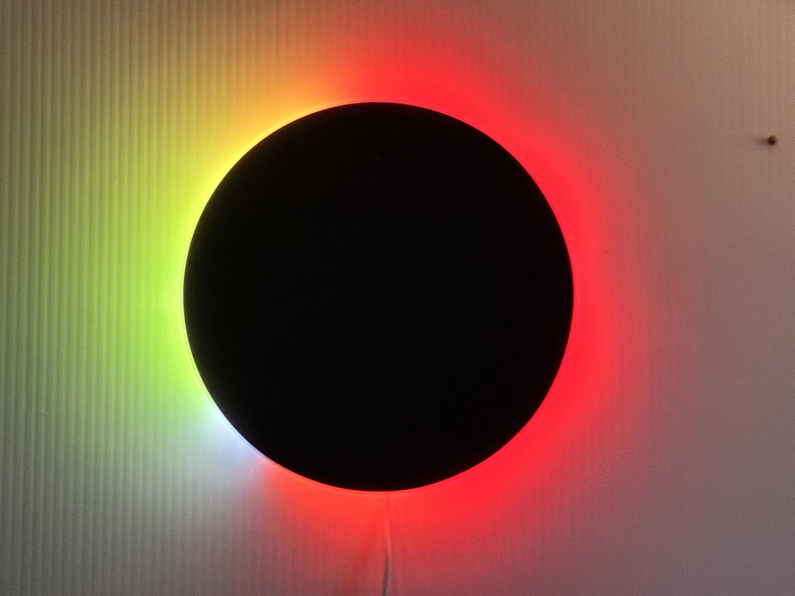 The image shows a circular black disc mounted on a vertically ribbed white wall. Surrounding the disc is a luminous halo with a gradient of colours transitioning from green on the left to yellow, then bright, angry red. A faint cord hangs down from the disc, suggesting it may be an illuminated wall fixture or art installation.