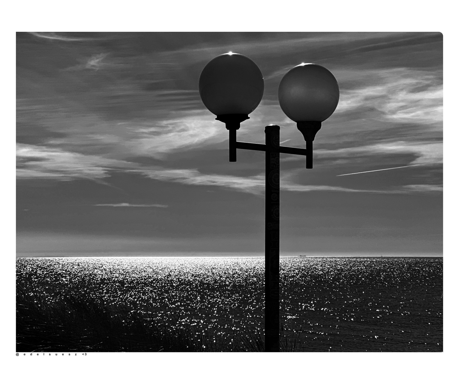 Aufnahme in schwarzweiß, Blick auf zwei ballonförmige Straßenlampen, befestigt an einem Laternenmast, im Hintergrund die dunkle Meeresoberfläche der Ostsee mit tausenden kleinen Lichtpunkten, Spiegelungen der Sonne, hellgrauer Himmel mit weißen Schleierwolken darüber