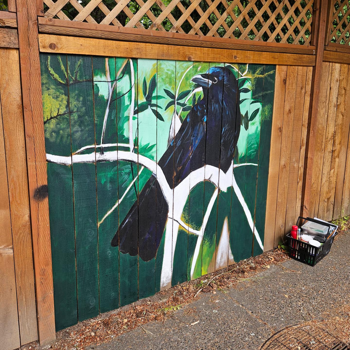 an 8ft by x6ft in progress mural on a cedar fence of a crow looking to the left on a birch branch amidst green leaves and mossy branches