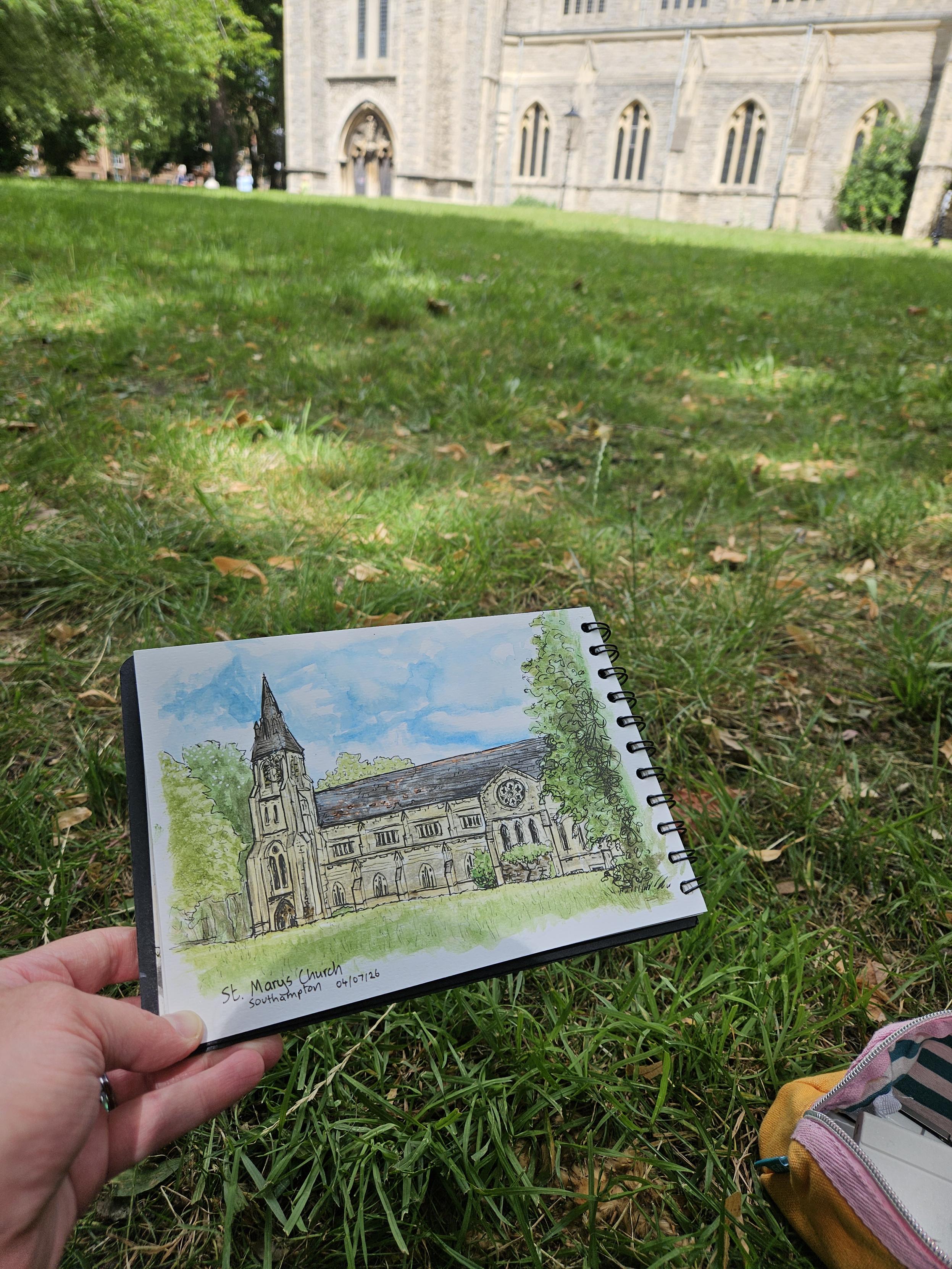 A sketchbook held beside some grass, with a watercolour mixed media sketch of a large church which can be partially seen in the background.
