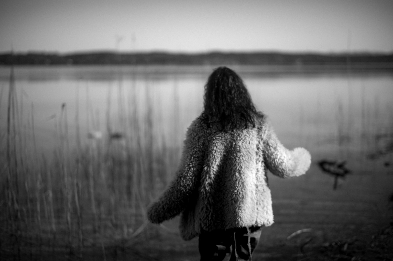 Eine Person mit lockigem Haar und einem flauschigen hellen Mantel steht mit dem Rücken zum Betrachter am Ufer eines ruhigen Sees. Schilf rahmt die Szene ein, während die Landschaft im Hintergrund durch die geringe Schärfentiefe weich verschwimmt. Die Schwarz-Weiß-Aufnahme vermittelt Ruhe, Nachdenklichkeit und Weite.