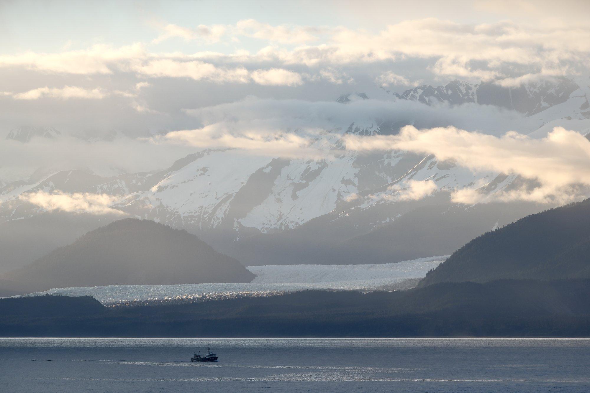 Hazy light illuminates fog and ice-capped mountains, with the edge of a glacier gleaming in the last vestiges of light. A ship travels across the water in front of it all.