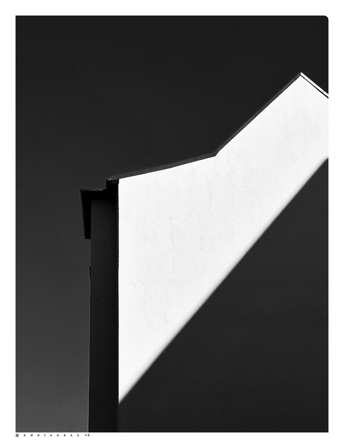 Aufnahme in schwarzweiß, Blick von unten auf eine seitliche spitz Zulauf Hauswand, hell und ohne Fenster, schräg über die Fläche ein dunkler Schatten eines anderen Gebäudes, links und darüber dunkler Himmel