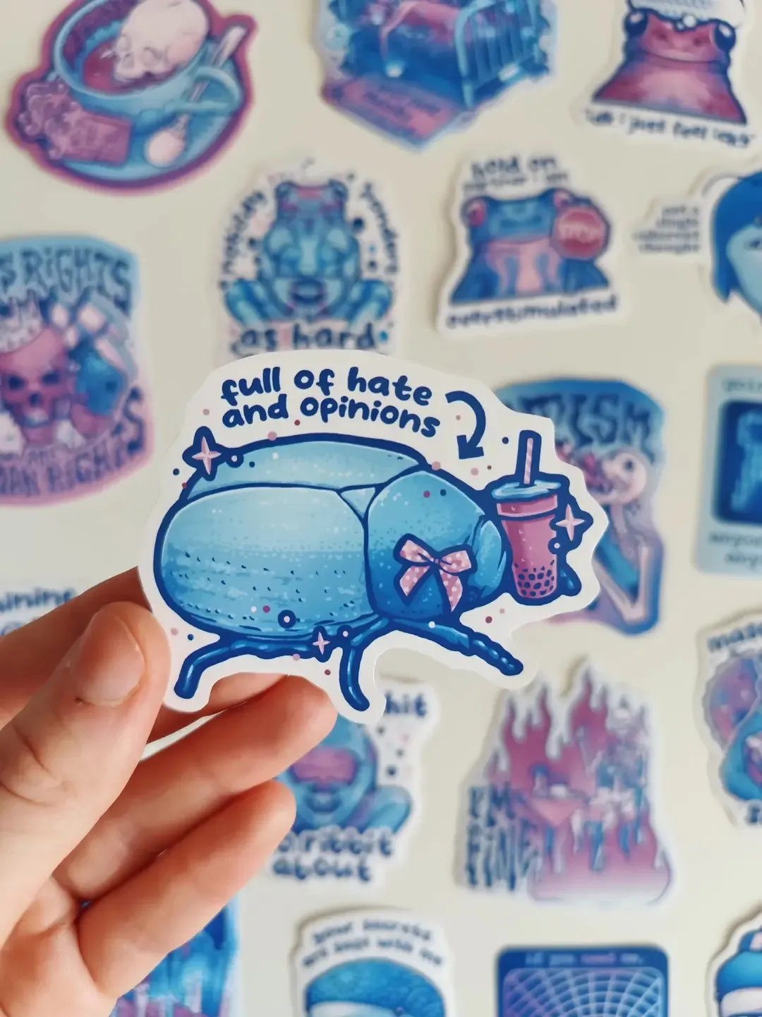 and holding up a sticker of a beetle with a boba tea titled "full of hate and opinions"