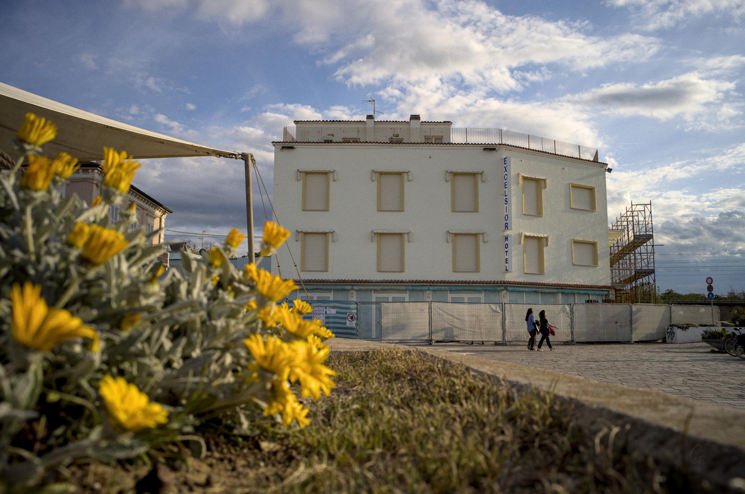A photo capturing an old, cheap hotel, under renovation. In front of it, people are passing by. Close to the camera, on the left, a bunch of yellow flowers appear gigantic compared to the far hotel.