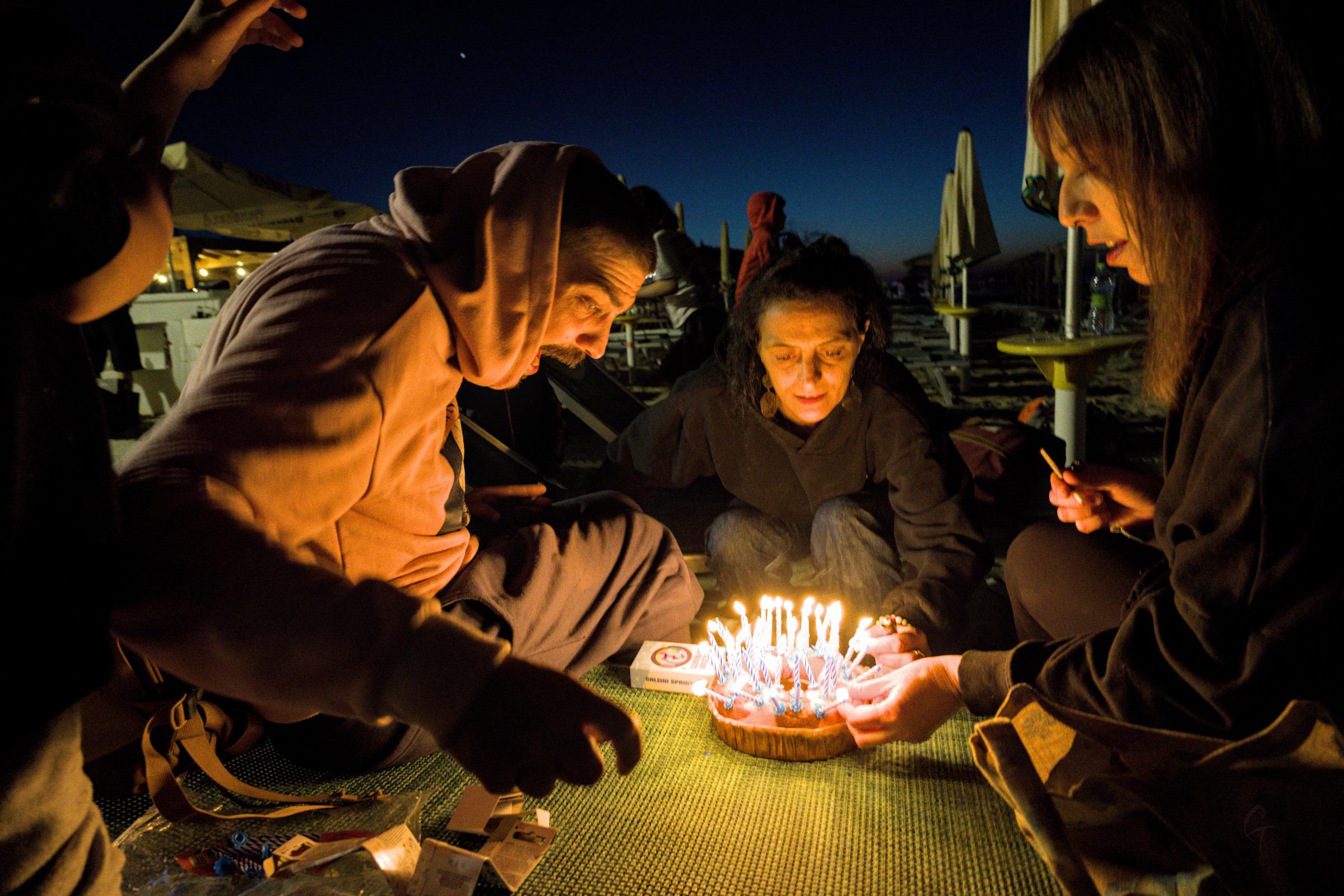 Three people are trying to light up many birthday candles that rest on a too-small cake in many different angles