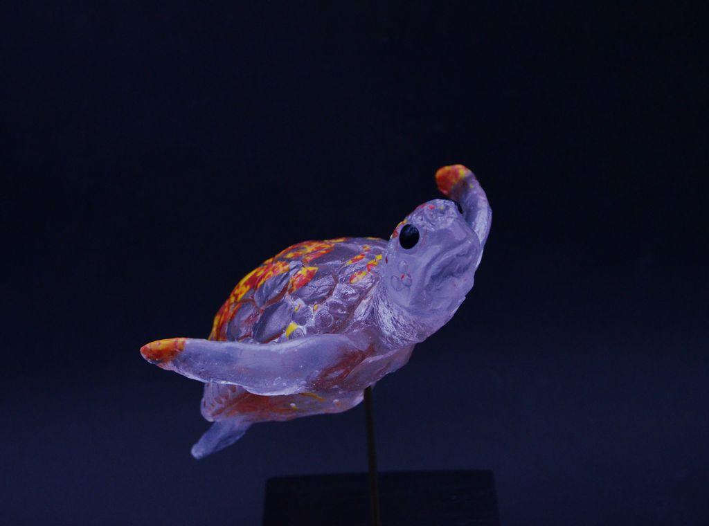 Photo d'une tortue en verre transparent et rouge/jaune pour le dos 
elle donne l'impression de nager
elle est sur un socle 
sur fond noir 
de 3/4