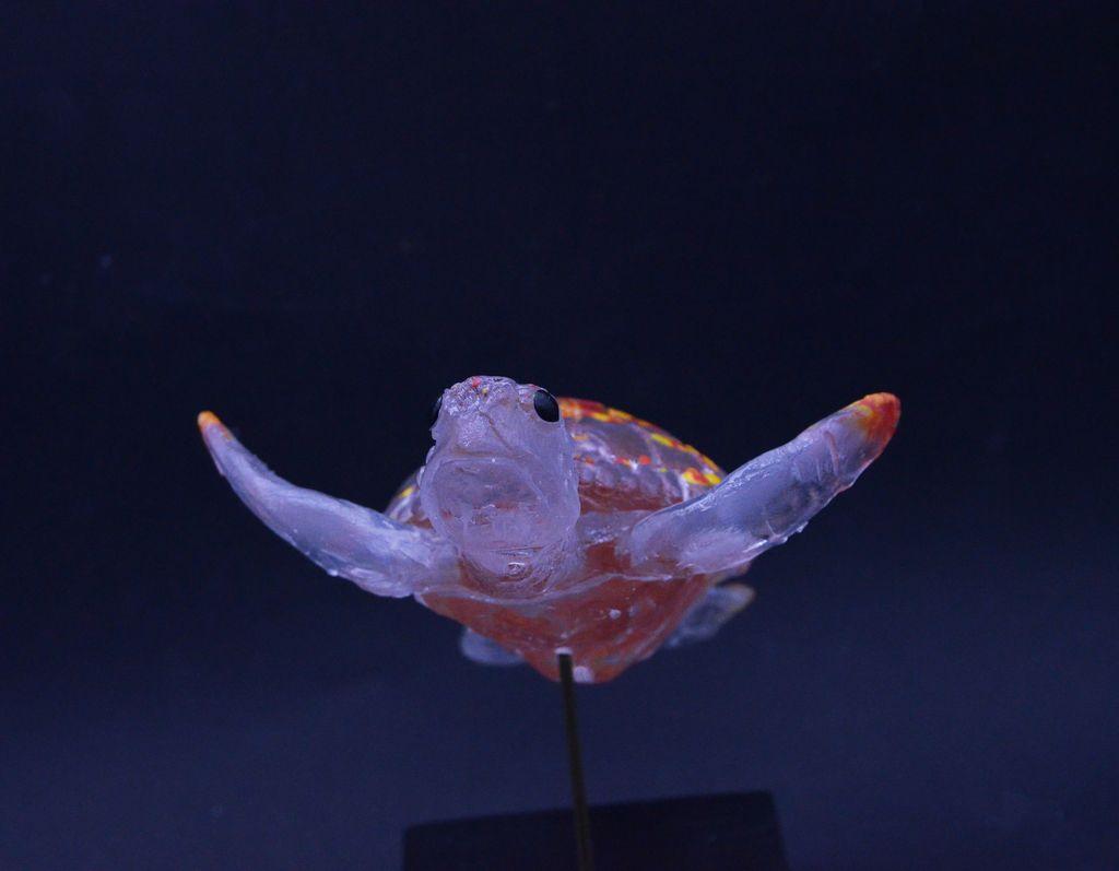 Photo d'une tortue en verre transparent et rouge/jaune pour le dos 
elle donne l'impression de nager
elle est sur un socle 
sur fond noir 
de face