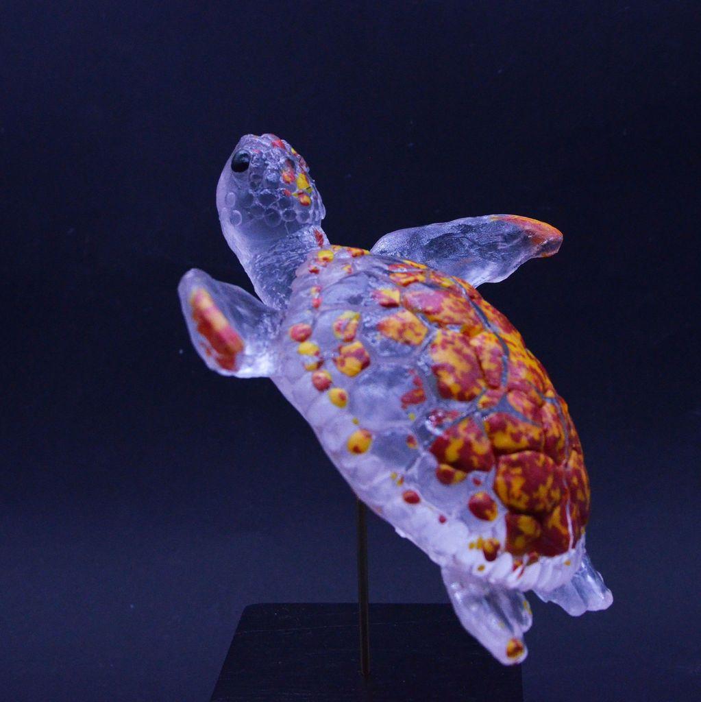 Photo d'une tortue en verre transparent et rouge/jaune pour le dos 
elle donne l'impression de nager
elle est sur un socle 
sur fond noir 
de profil