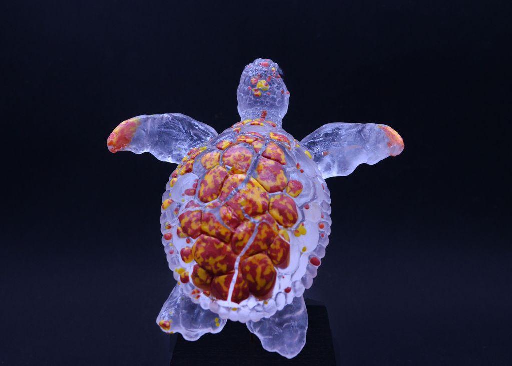 Photo d'une tortue en verre transparent et rouge/jaune pour le dos 
elle donne l'impression de nager
elle est sur un socle 
sur fond noir 
de dos