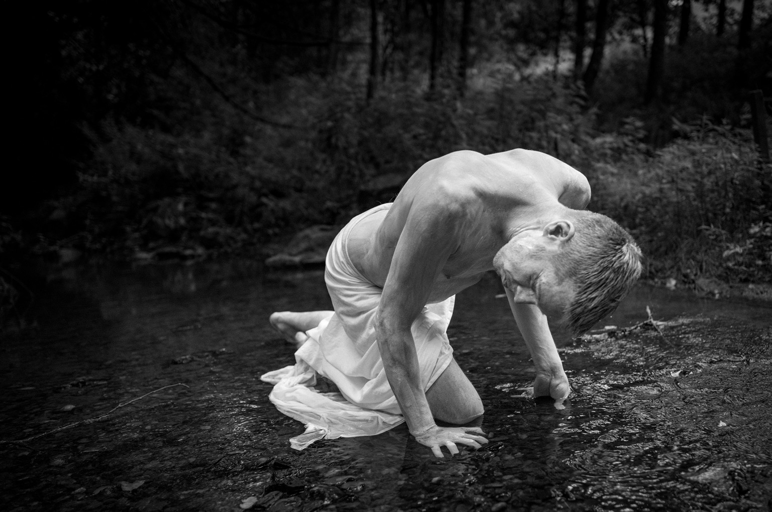 Ein Butoh-Tänzer kniet mit tief gebeugtem Oberkörper in einem flachen Waldbach. Seine Hände stützen sich im Wasser ab, ein weißes Tuch umhüllt seine Hüfte. Die angespannte Körperhaltung und die dunkle Waldkulisse verleihen dem Schwarzweißfoto eine intensive, schmerzhafte und fast mythische Atmosphäre.
