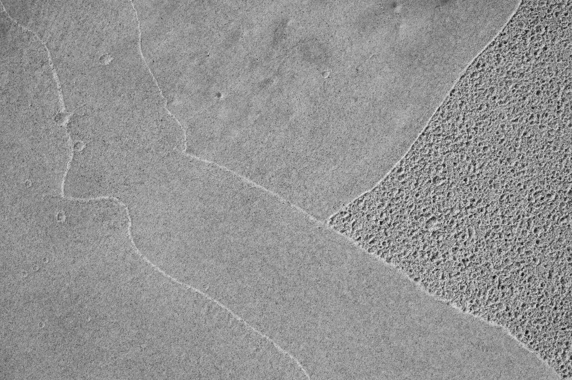 black and white photo of the sand with lines leftover from the wave edges. One triangular section of the sand is dotted after the rain, others are clean.