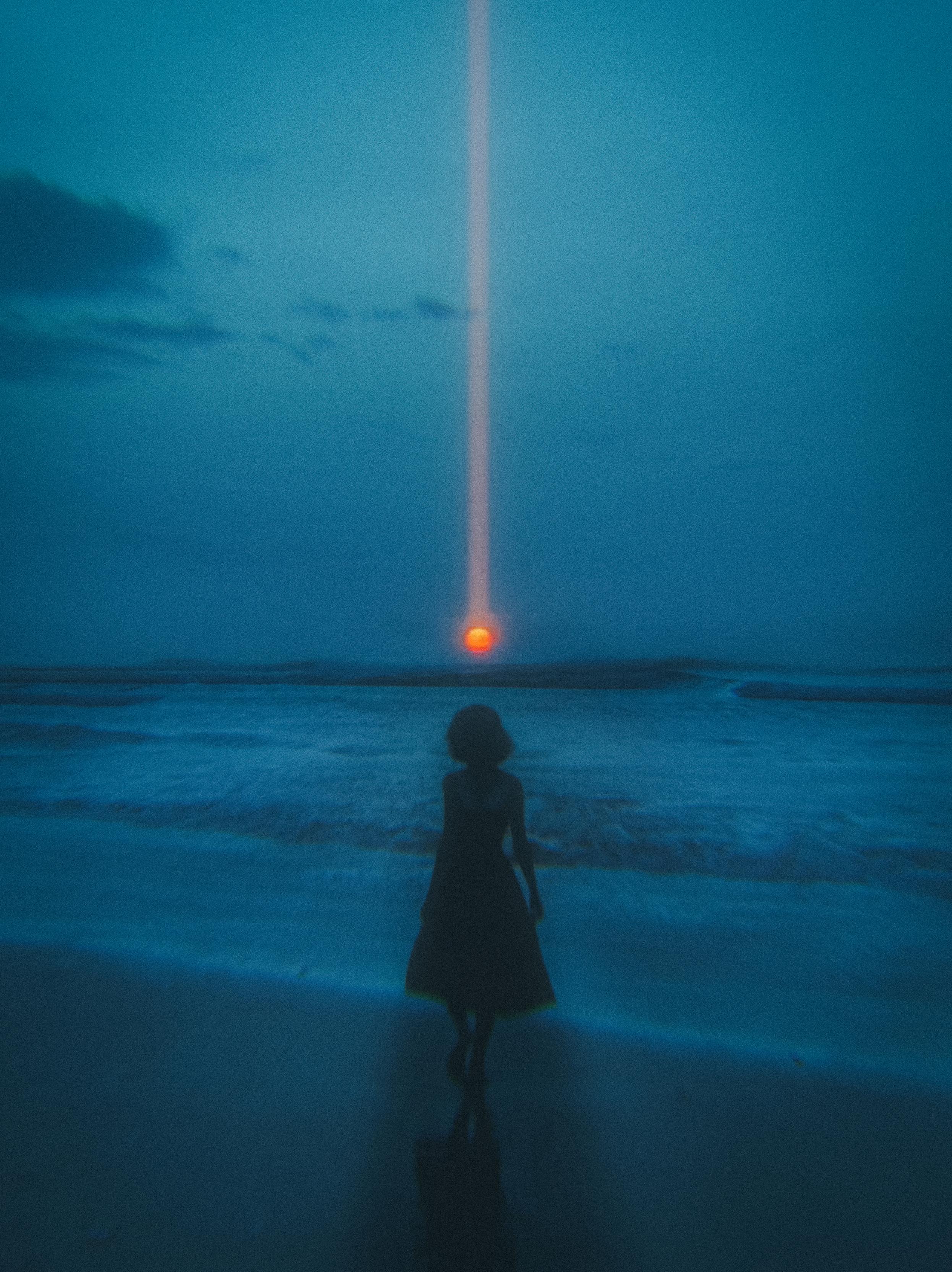 Digital art titled "New Dawn": A silhouetted figure in a vintage dress stands on a dark, wet shoreline facing a deep-blue ocean at twilight. On the horizon, a glowing orange sun emits a striking, vertical beam of warm amber light that stretches straight up into the dark blue sky.