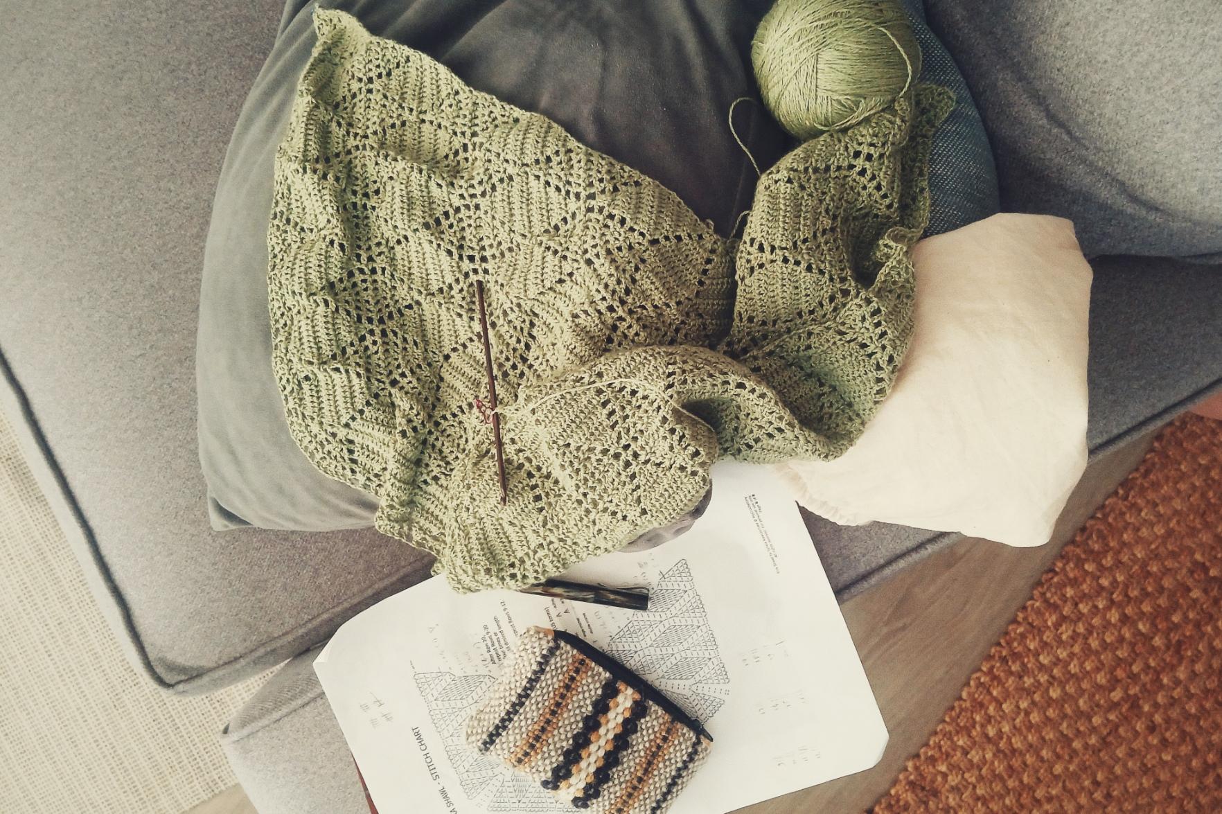 A vertical colour photograph of a green crochet shawl in progress. It rests on a grey sofa alongside the chart, pencil, and pouch to hold crochet bits and bobs.