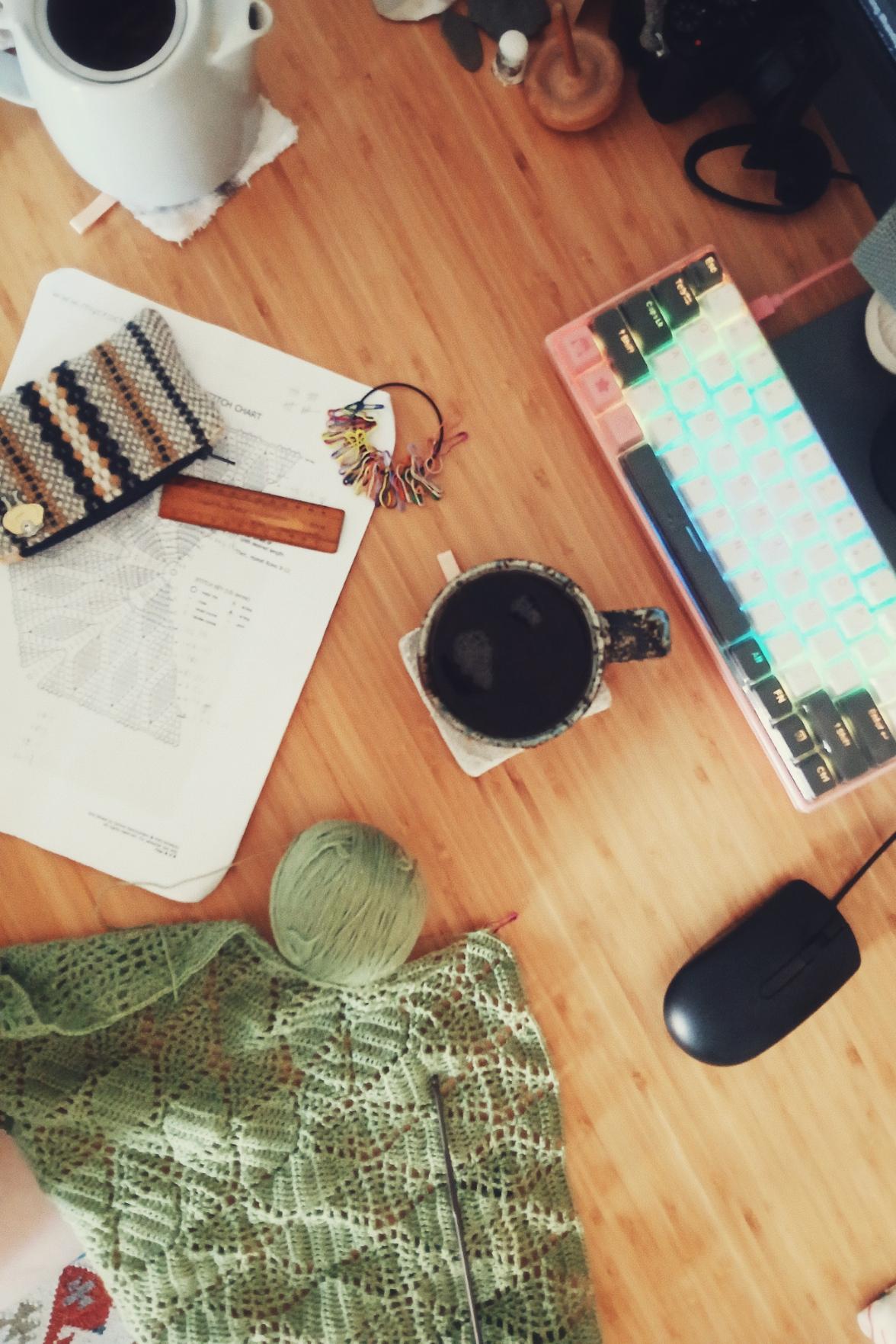 A vertical colour photograph of a green crochet shawl in progress on a desk. It is surrounded by tea, the chart, crochet bits and bobs, a lit keyboard, and a black mouse.