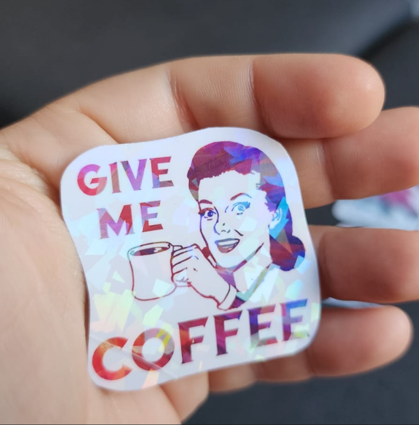 A holographic sticker that reads 'Give Me Coffee' with a vintage illustration of a woman holding up a cup of coffee